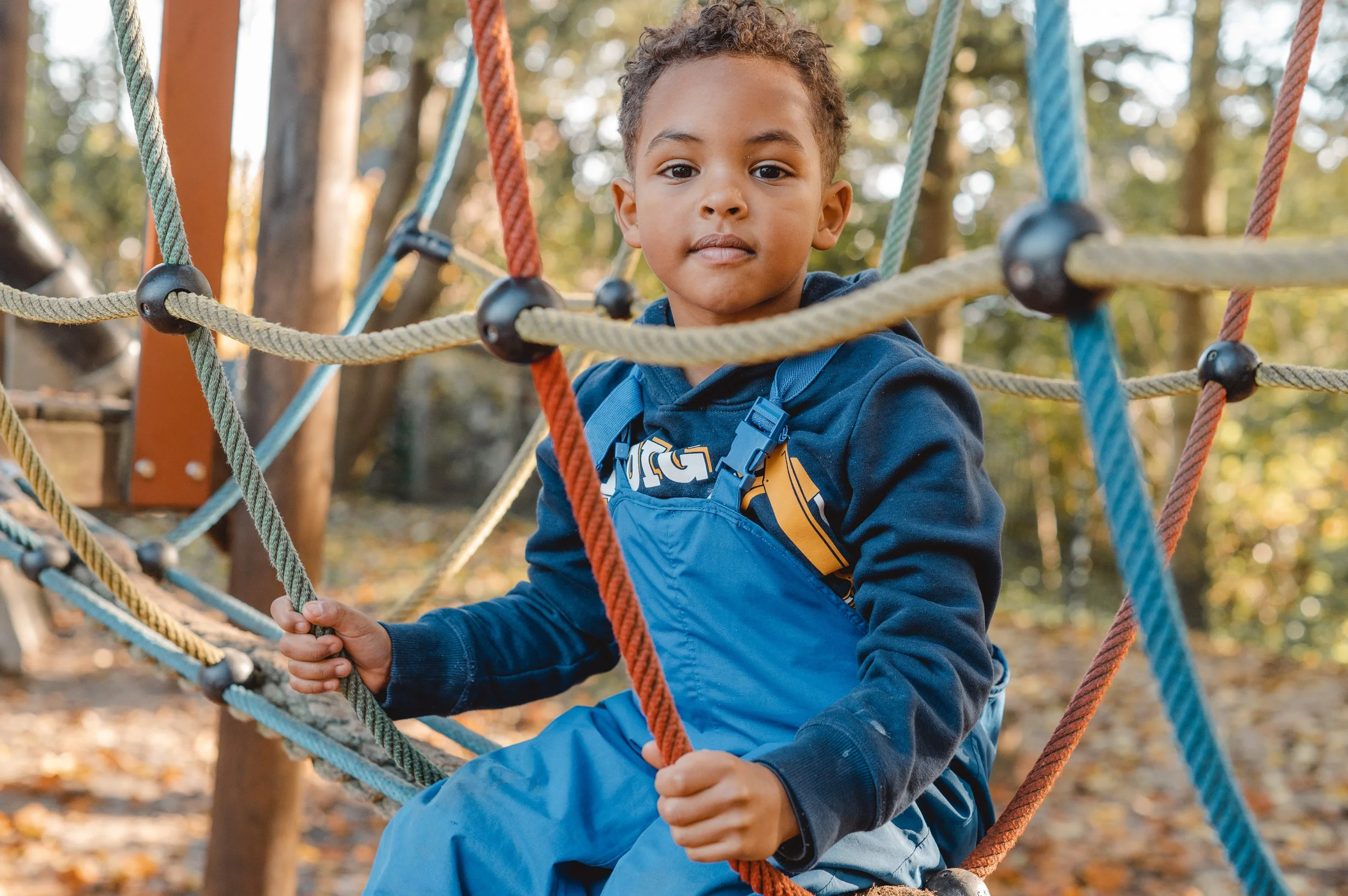 Ein Junge sitzt auf einer Kletterbrücke aus Seilen, umgeben von Bäumen und Herbstblättern.