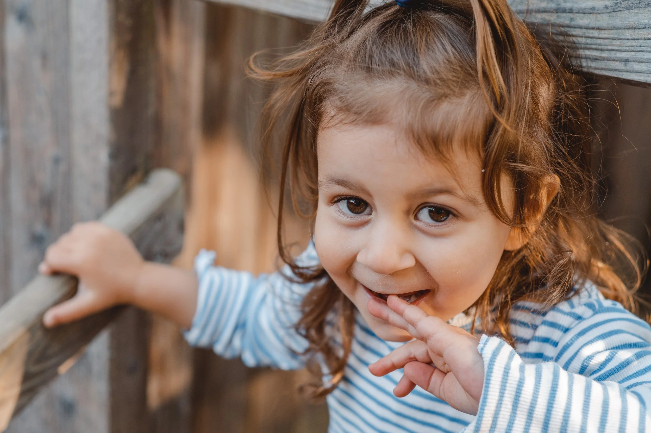 Ein Mädchen mit braunen Locken trägt einen gestreiften blauen und weißen Pullover, hält sich an einem Holzgeländer fest mit ihrem Finger am Mund, während sie in die Kamera lächelt.