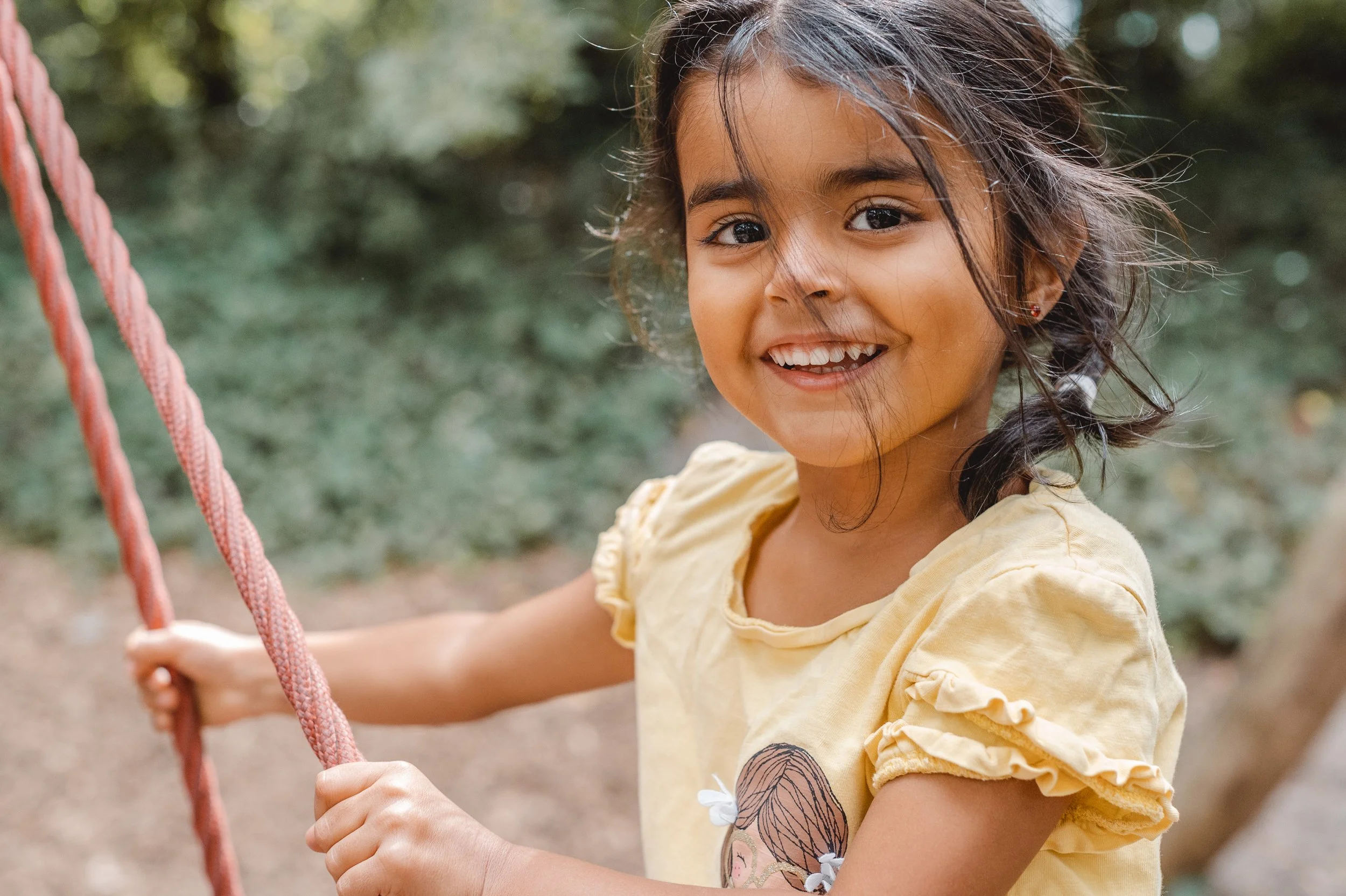 Lächelndes Mädchen im gelben T-Shirt auf einer Schaukel im Freien, umgeben von Bäumen.