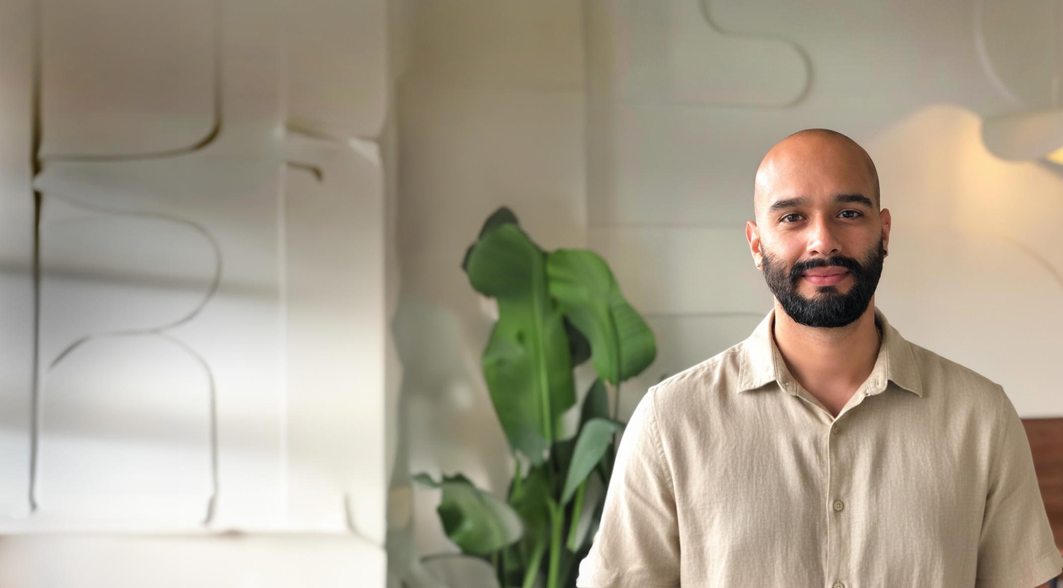 A man with a bald head and beard, wearing a beige shirt, standing indoors near a large leafy plant and a modern white wall with abstract line art.