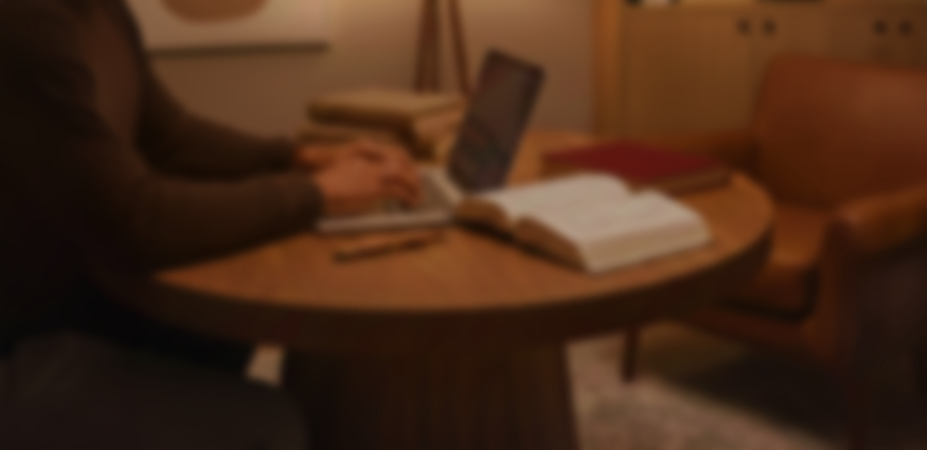 A person working on a laptop at a round wooden table with an open book and a closed book on it, in a cozy, warmly lit room.