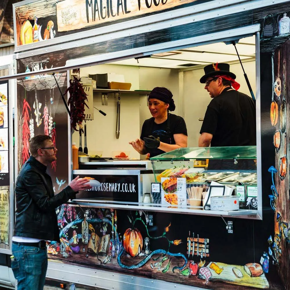 A food truck serving Mexican food, with two staff members preparing food and a customer ordering at the window. The truck has colorful artwork depicting pumpkins, bread, and kitchen items.