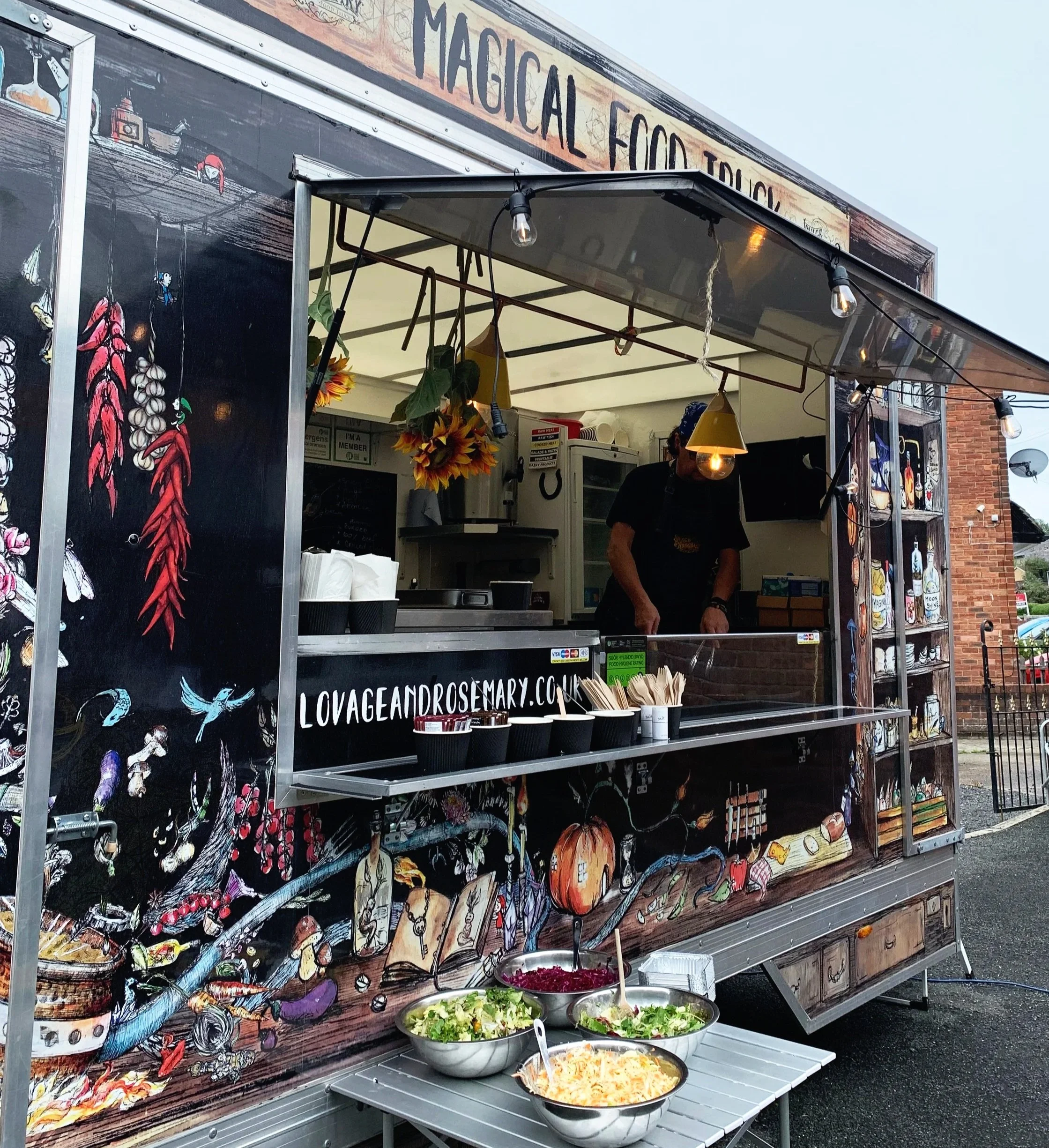 Lovage and Rosemary food truck decorated with colorful illustrations of food and vegetables, serving salads and other dishes with bowls of salad in front.