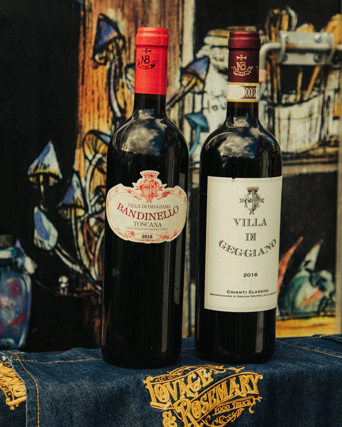 Two bottles of red wine stand on a table with a dark cloth, featuring gold embroidery. The background is a colorful, abstract painting.