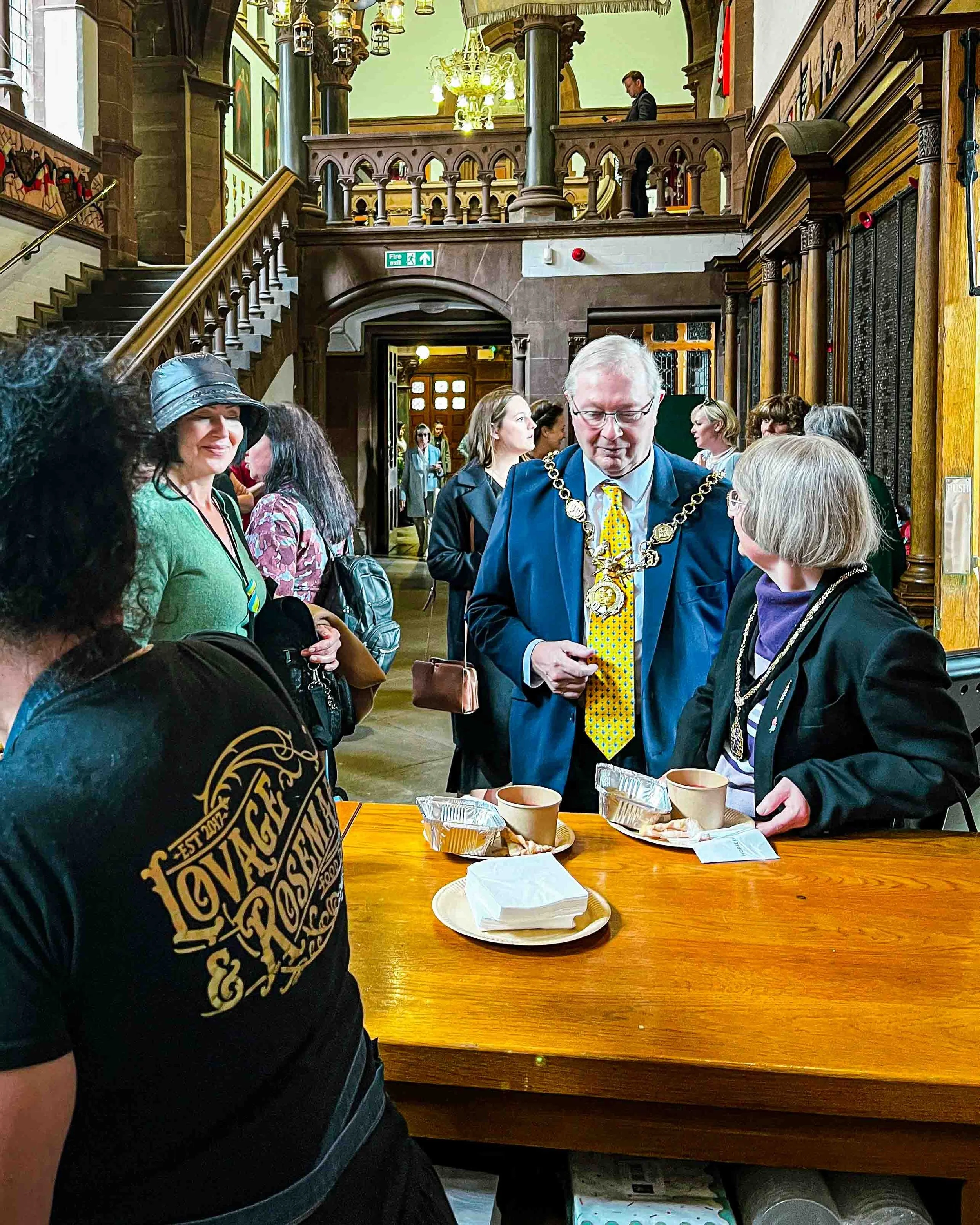 A group of people gathered in a historic, ornate room with wooden architecture, chandeliers, and portraits on the walls. One man is wearing a chain of office, suggesting a formal or official event.