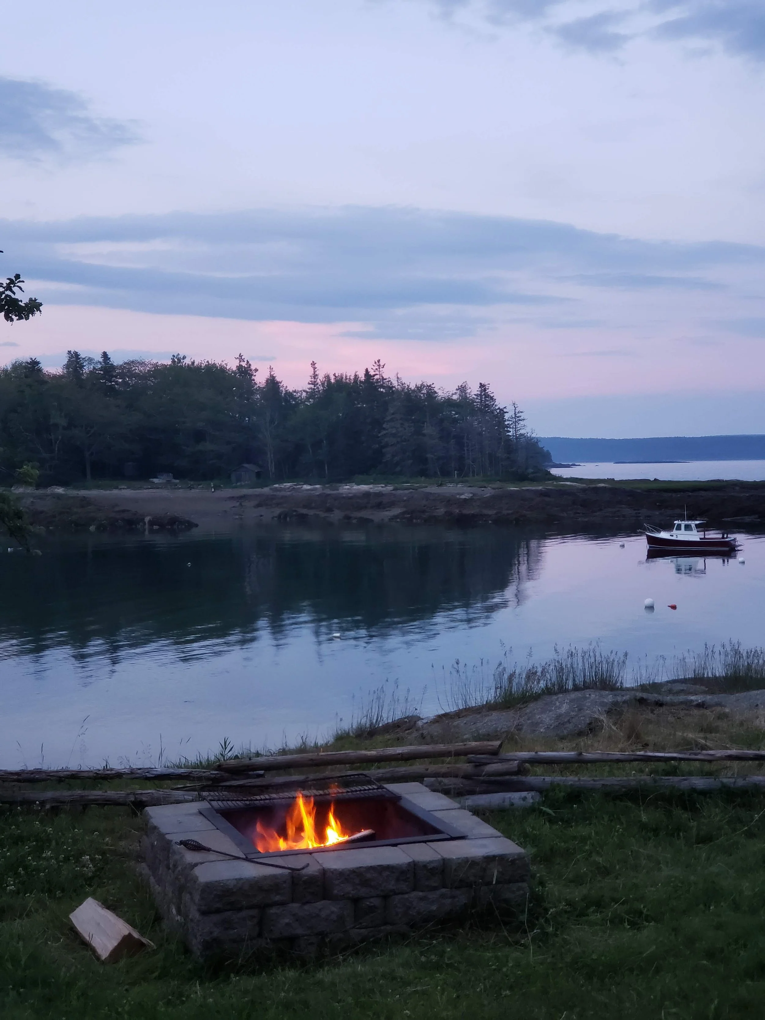 A waterfront scene at dusk or dawn with a small fire in a stone firepit in the foreground, calm water reflecting the sky, a boat anchored near the shore, and trees along the distant shoreline.
