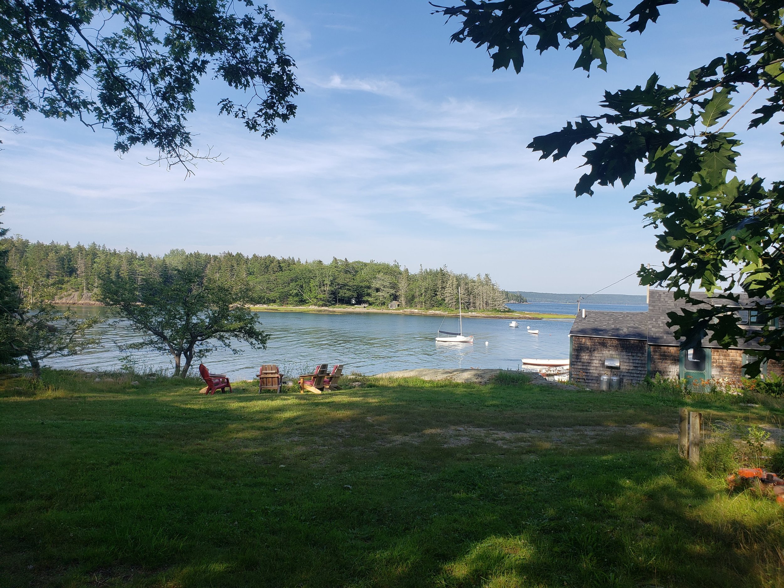 A peaceful lakeside scene with green grass, trees, Adirondack chairs, and a small house near the water. The lake has a few sailboats and boats anchored, with a forested shoreline in the background under a partly cloudy sky.