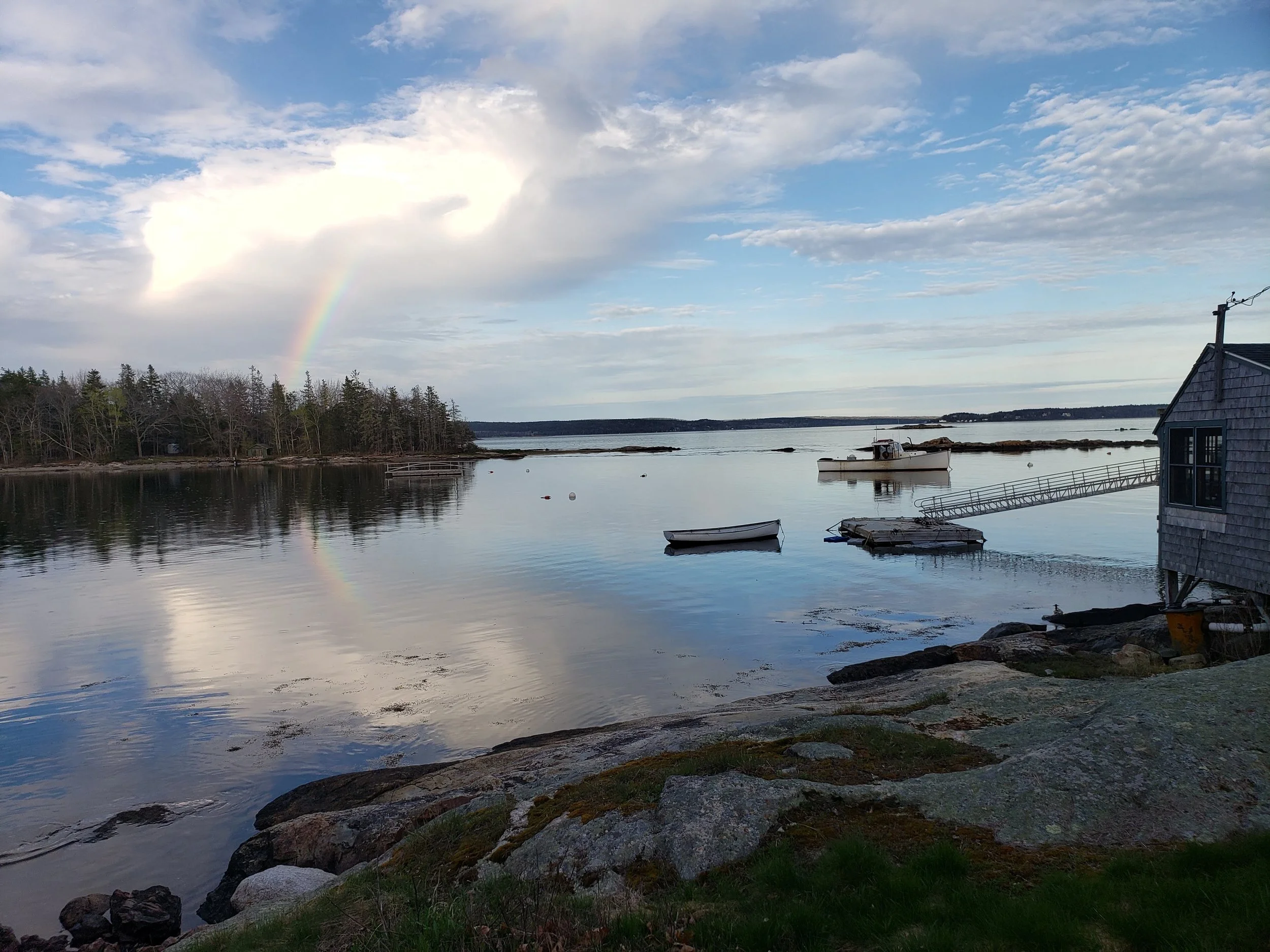 Seaside view with calm water, small boats, a wooden dock, and a house on the right, under a partly cloudy sky with a rainbow.
