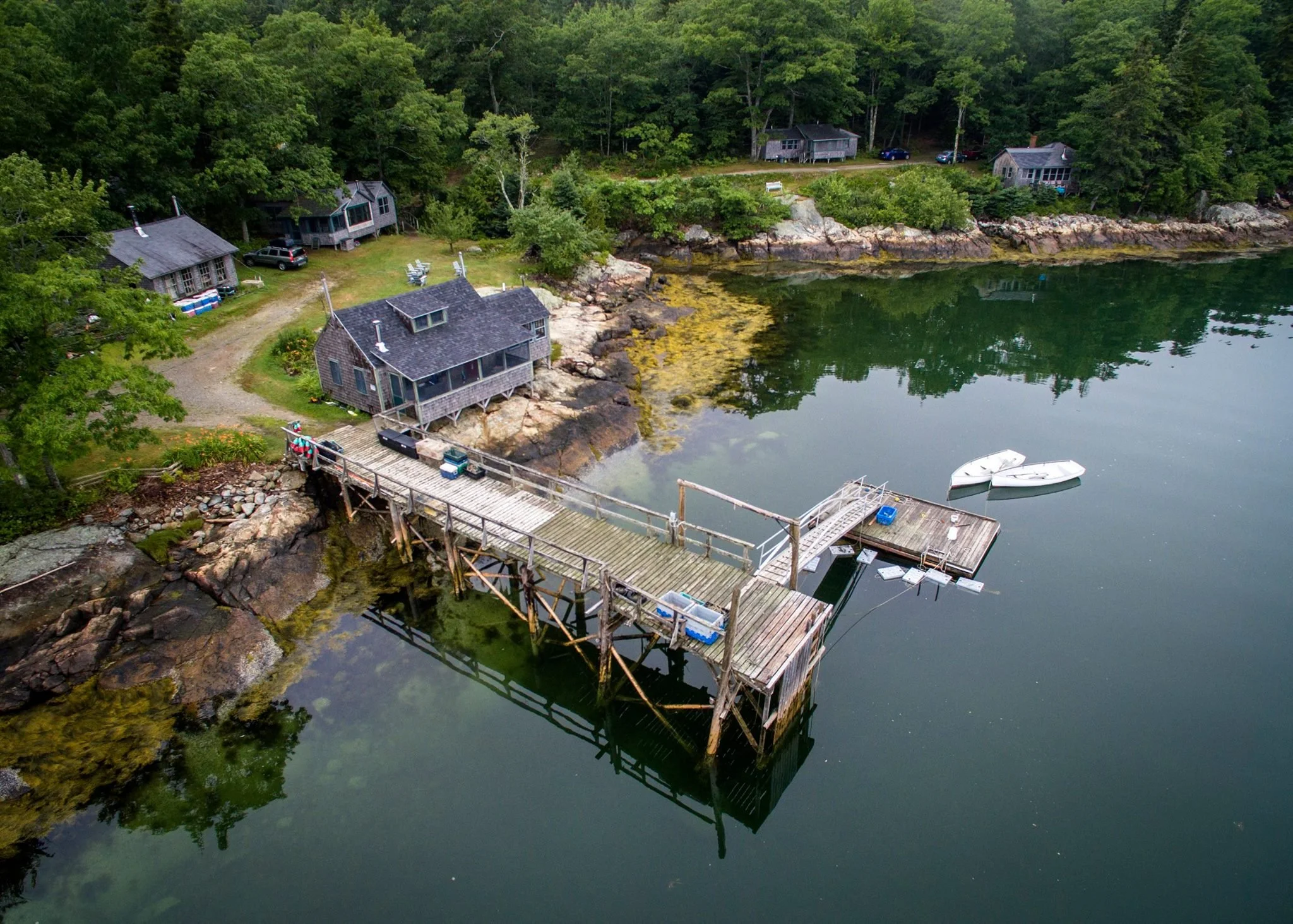 Aerial view of a lakeside cabin with a wooden dock extending into the water, boats moored nearby, and surrounding greenery with other cabins and trees.