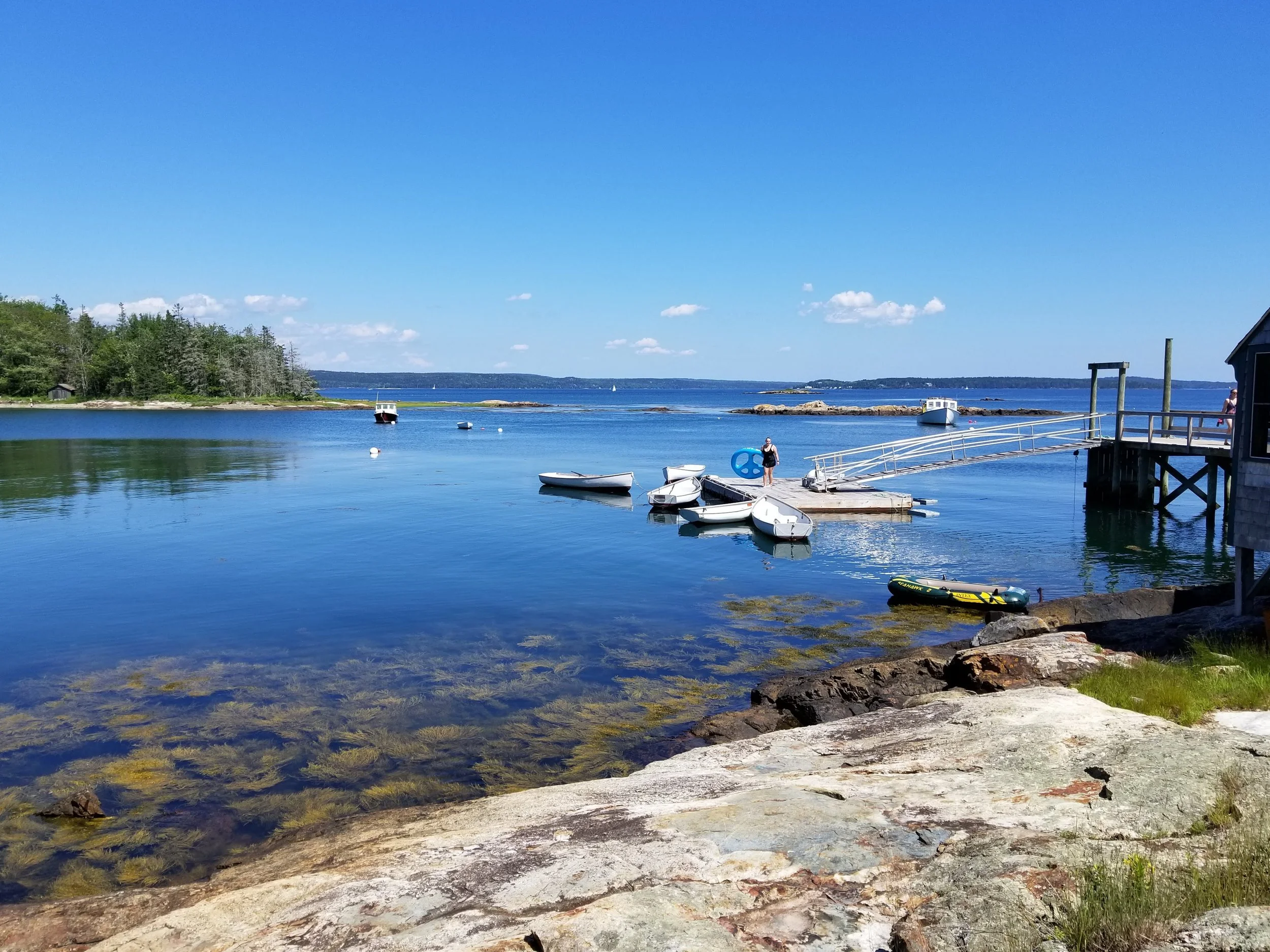 A peaceful waterfront scene with small boats docked near a rocky shoreline, a person standing on a floating dock, and a clear blue sky with scattered clouds.