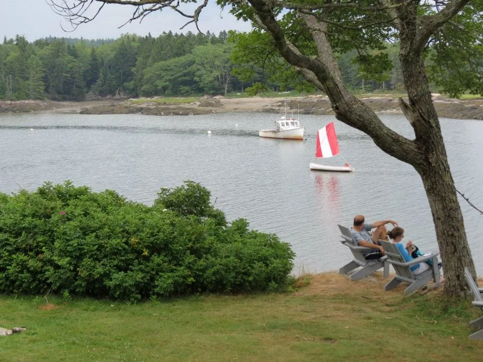 A couple sitting on Adirondack chairs under a tree by the water, overlooking a lake with sailboats, one with a red sail and one with a white sail, surrounded by greenery and hills.