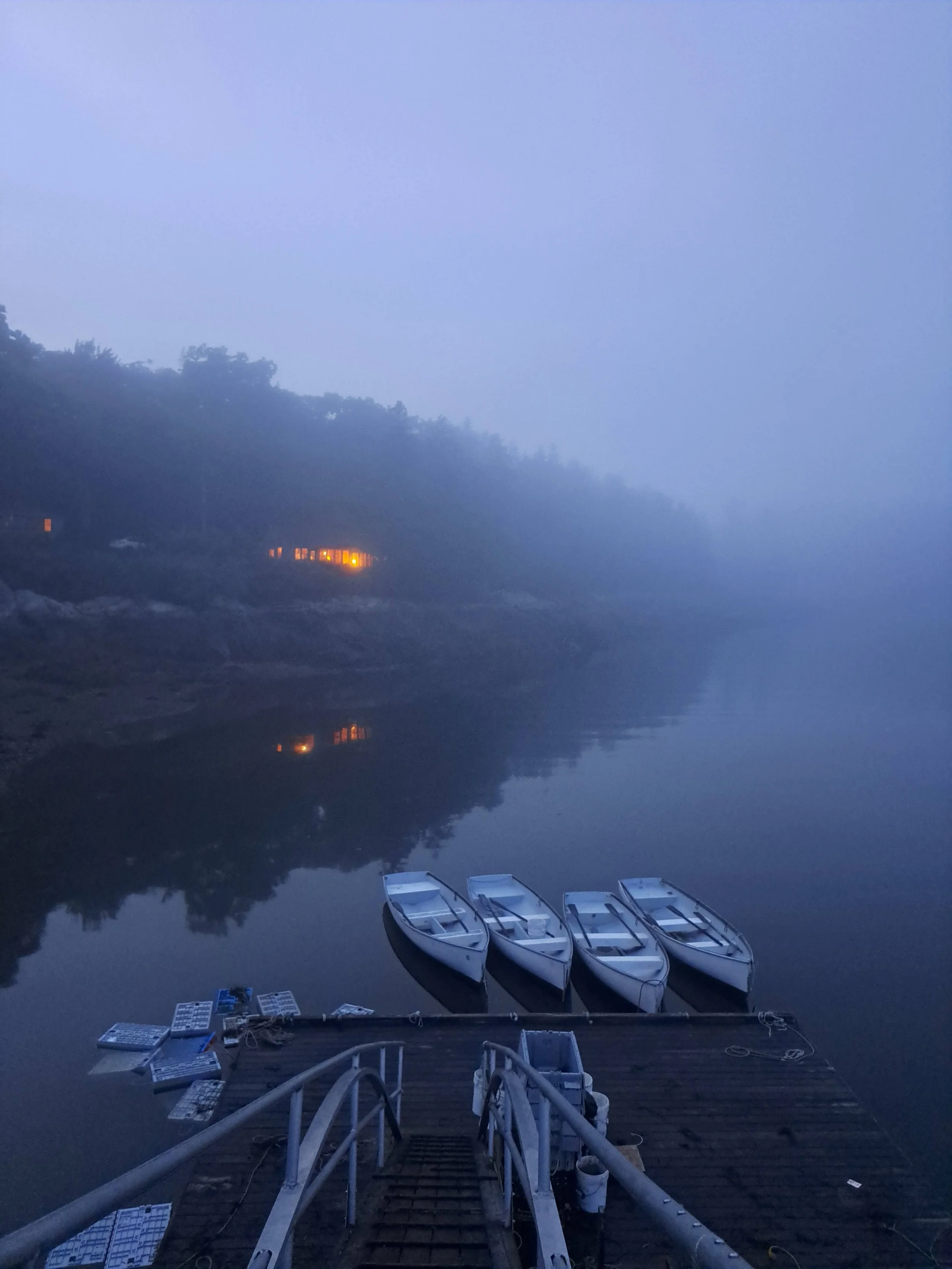 Four white boats moored at a wooden dock on a calm, foggy river at dawn, with faint lights on the shore and trees reflected in the water.