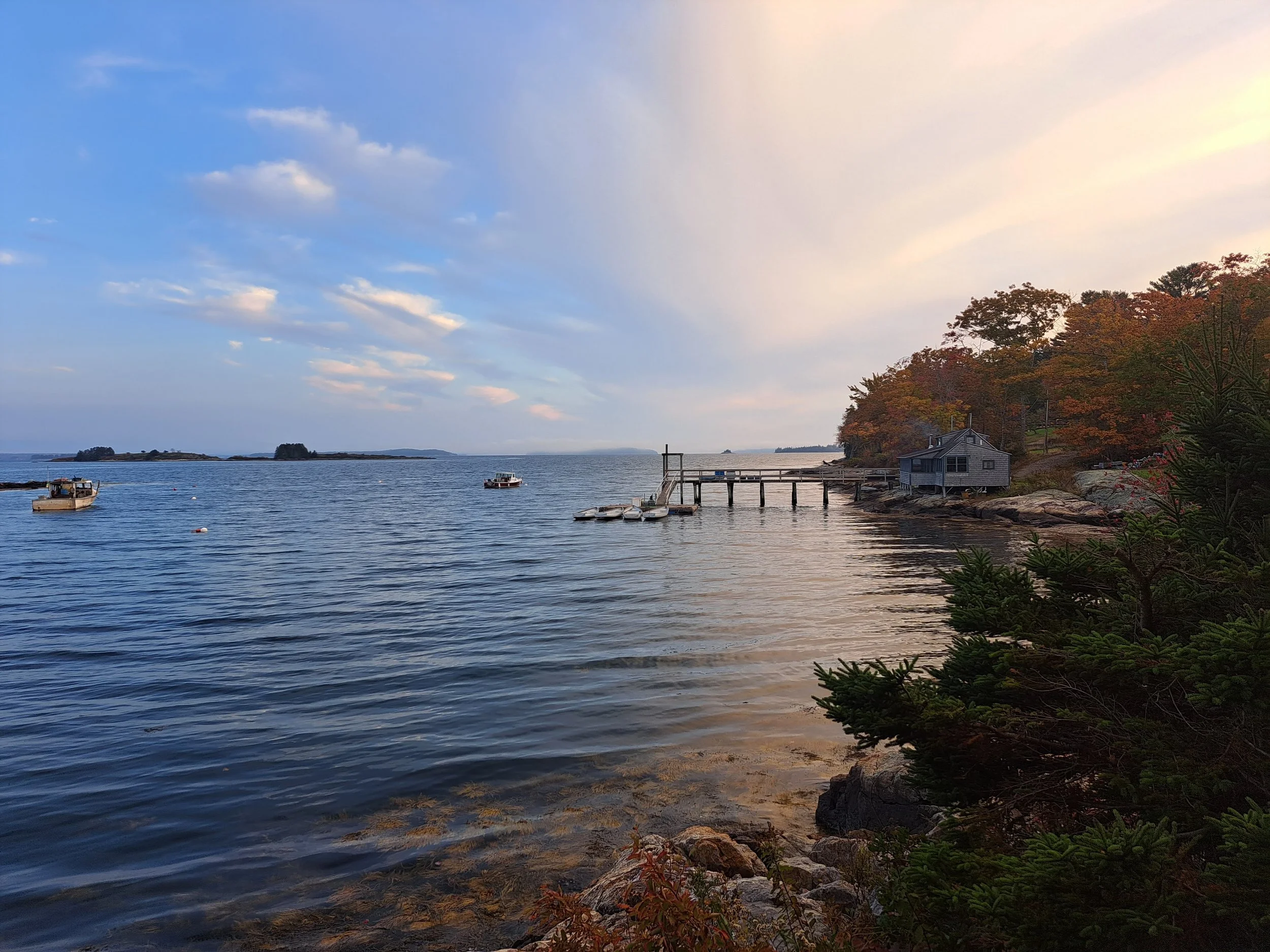 A tranquil coastal scene with boats floating on calm water, a small pier extending into the water, and a house near a shoreline surrounded by trees with autumn foliage. The sky has a mix of clouds and clear patches, indicating a peaceful evening or m
