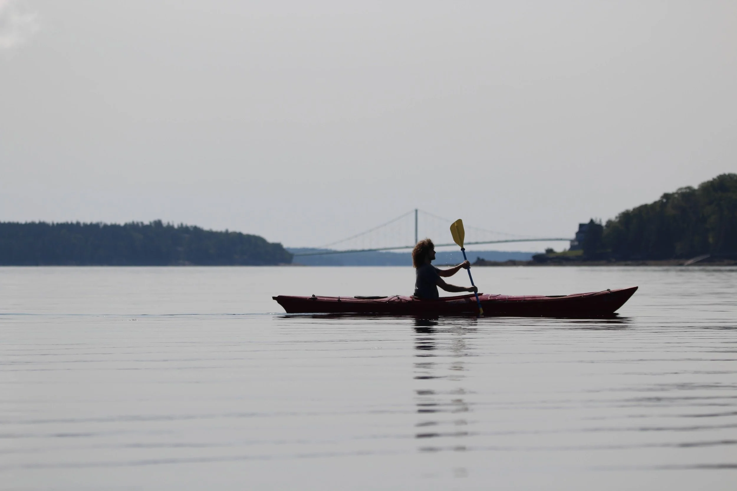 A person kayaking on a calm body of water with a bridge and land in the background.