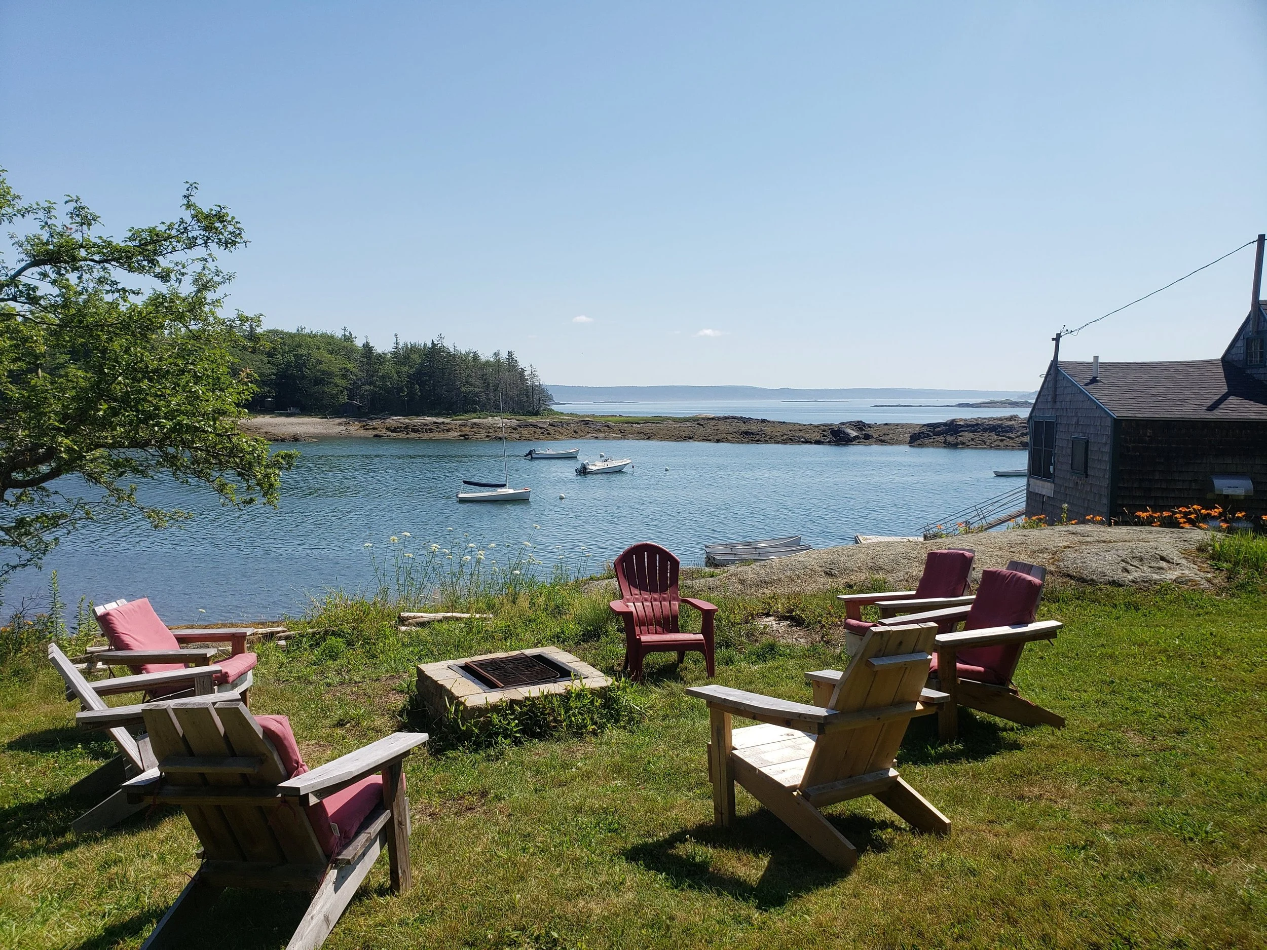 Seaside scene with five wooden chairs with pink cushions arranged around a small fire pit on a grassy area overlooking a calm body of water with sailboats, rocky shores, and a house with dark shingles.
