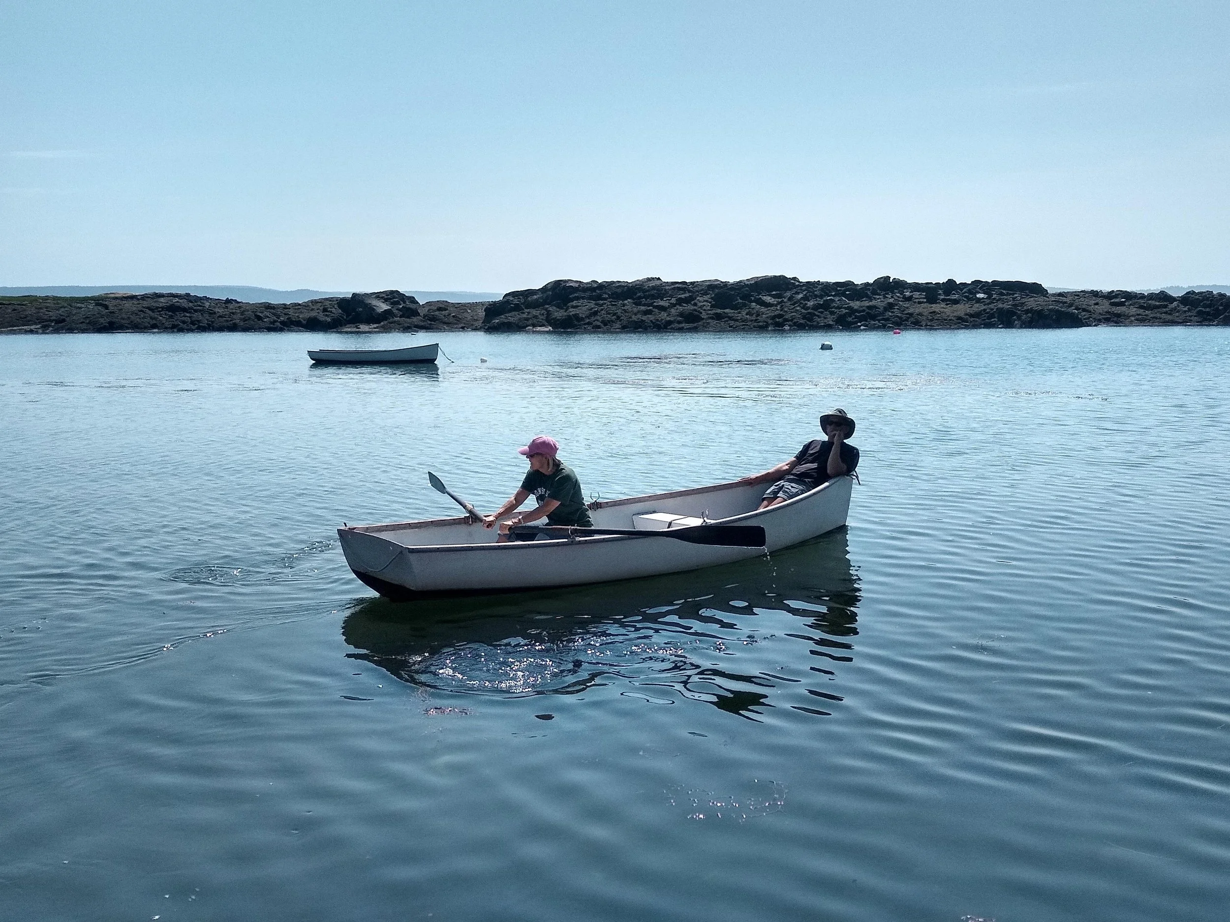 Two people in a small white boat on calm water, with a rocky outcrop and another boat in the background, under a clear blue sky.