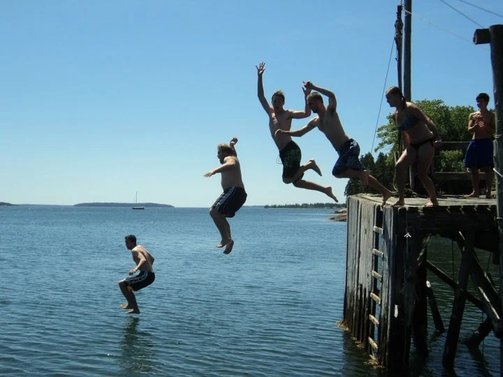 Group of children jumping off a wooden dock into a body of water on a sunny day.