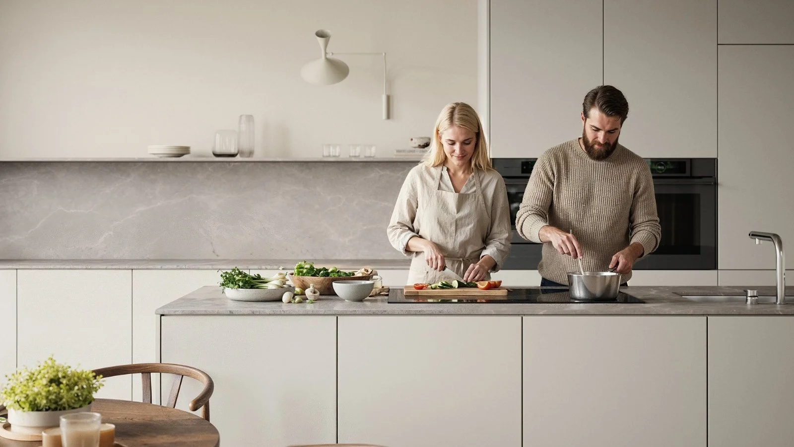 A man and a woman preparing food together in a modern kitchen with white cabinets and a marble backsplash, surrounded by vegetables and dishes on the counter.