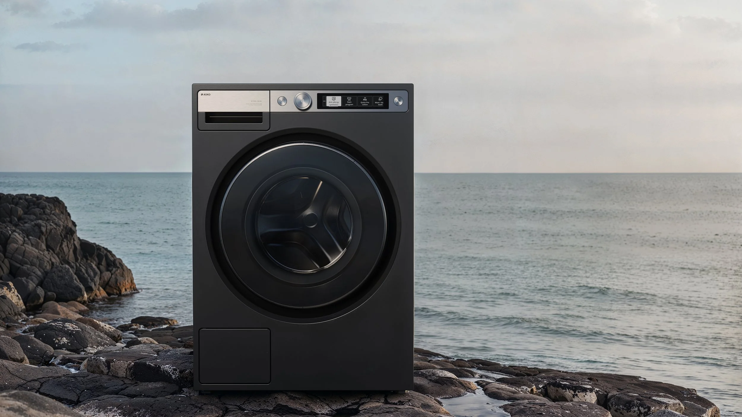 A black front-loading washing machine on a rocky shoreline by the ocean with cloudy sky.