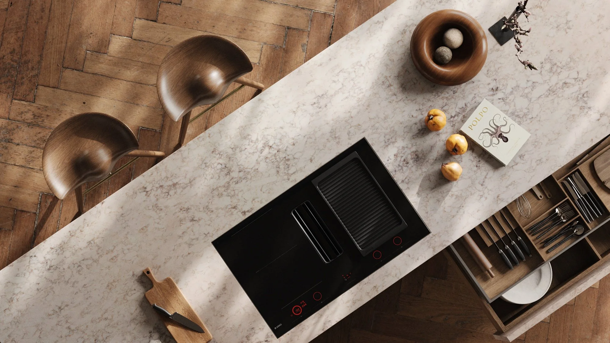 A kitchen countertop with a black induction cooktop, a wooden cutting board with a knife, a round wooden bowl with three decorative stones, a vase with branches, a box titled 'Polpo' with an illustration of an octopus, three golden-feathered jewelry 