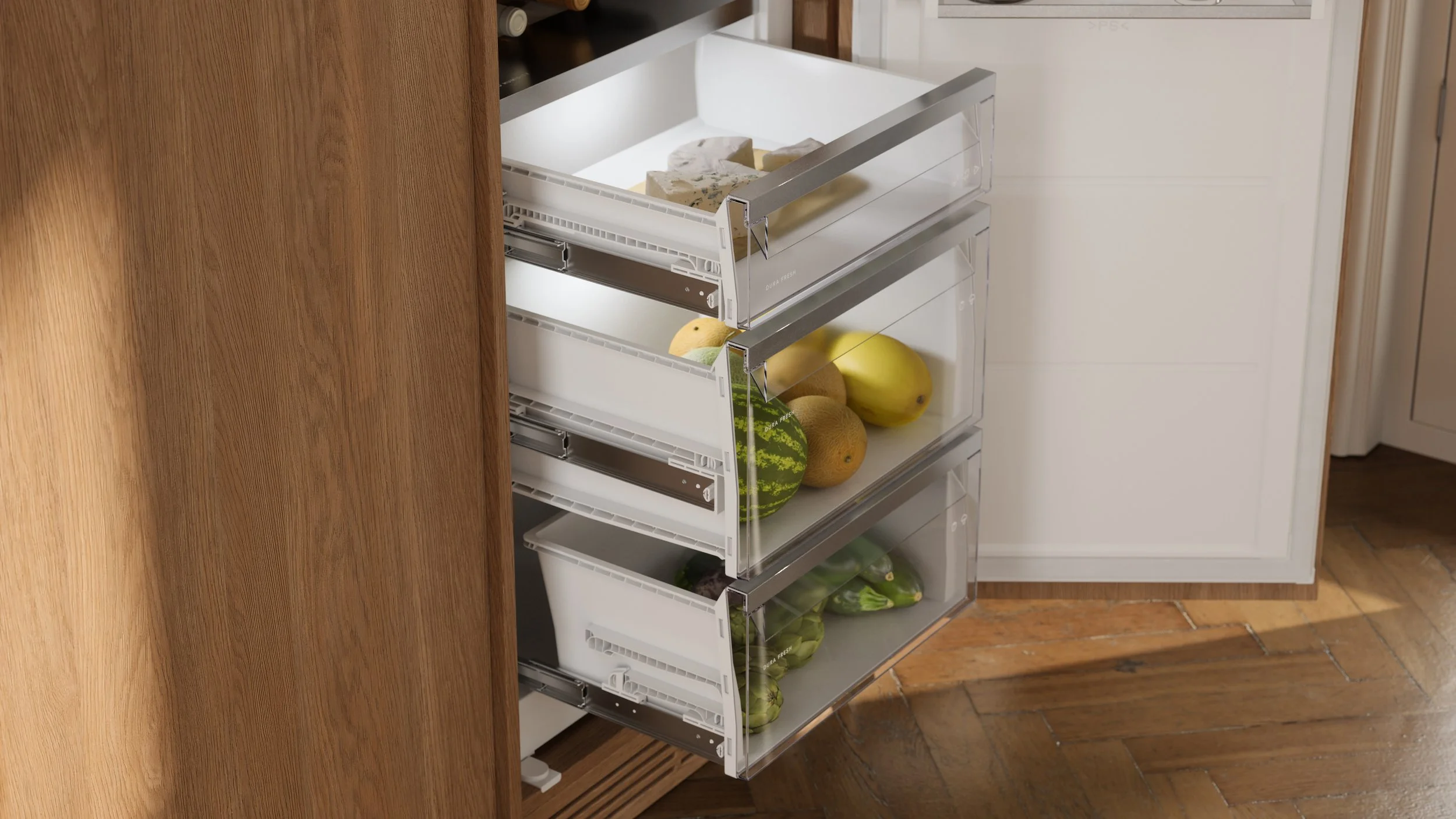 Open fridge drawer with various fruits and vegetables, including watermelon, cantaloupe, bananas, and cucumbers, next to wooden kitchen cabinets and hardwood floor.