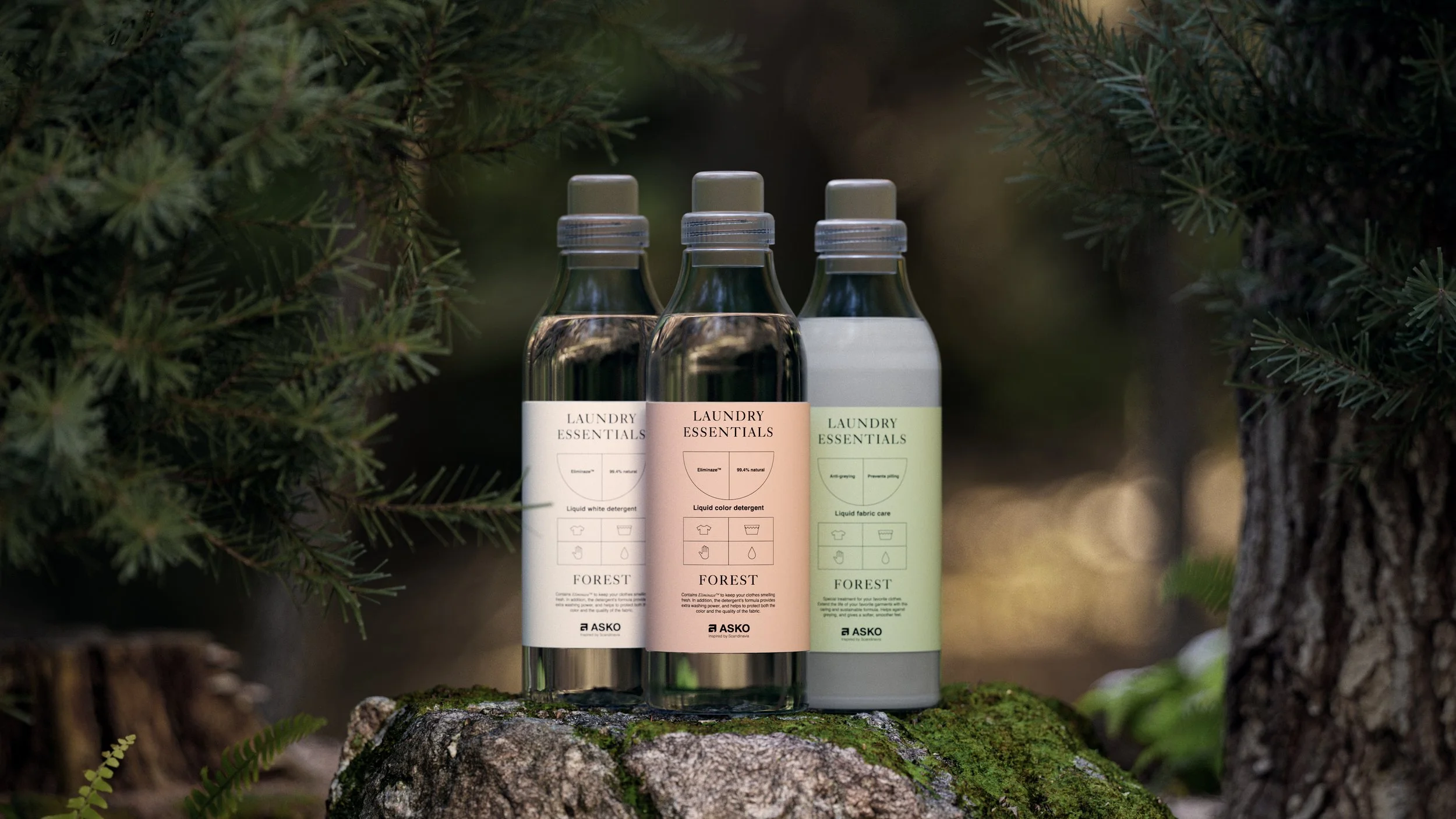 Three bottles of laundry detergents labeled 'Laundry Essentials,' placed on a moss-covered log outdoors surrounded by green pine trees.
