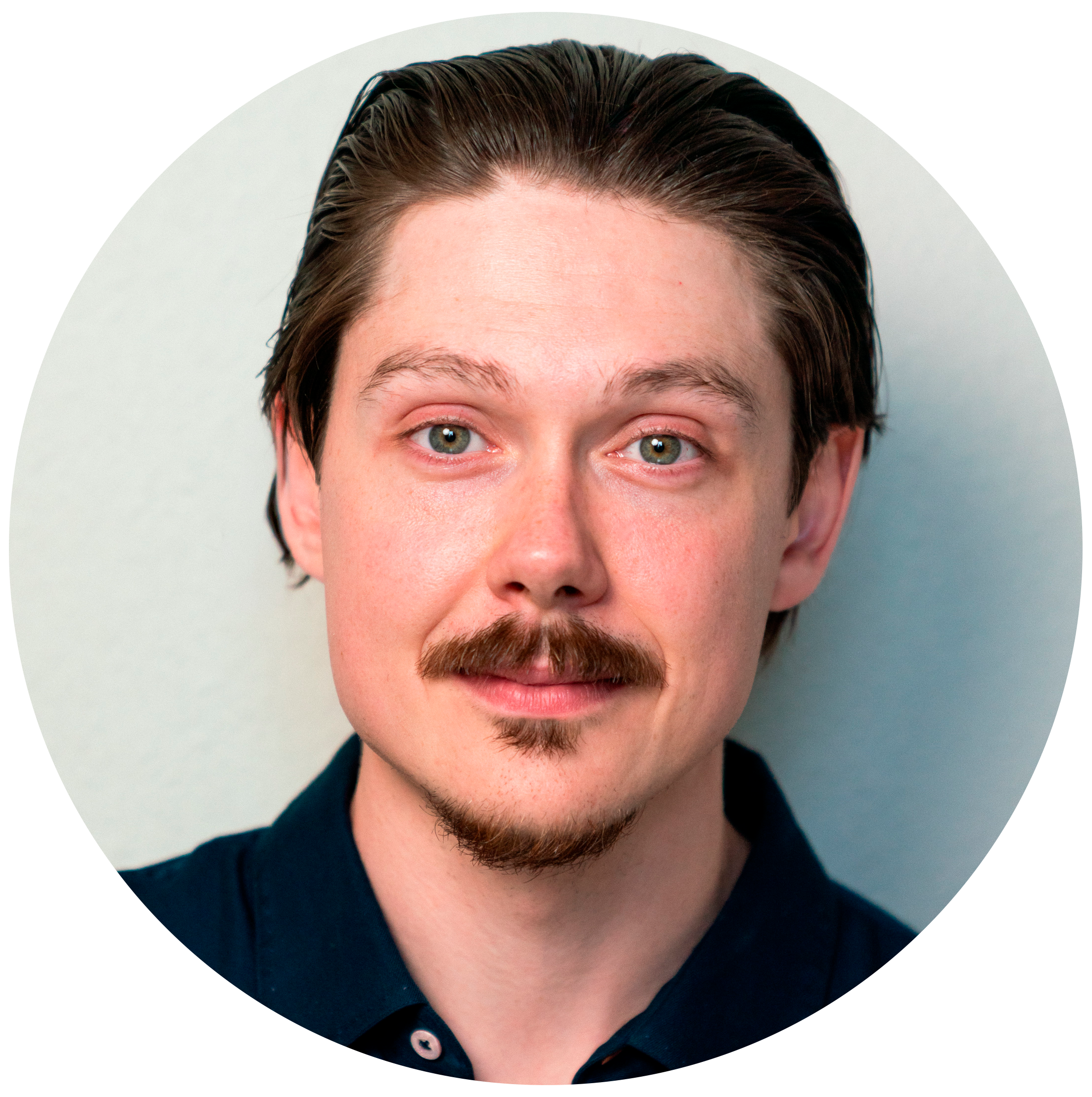 A young man with light skin, blue eyes, a mustache, and a goatee, wearing a dark shirt, against a plain light background.
