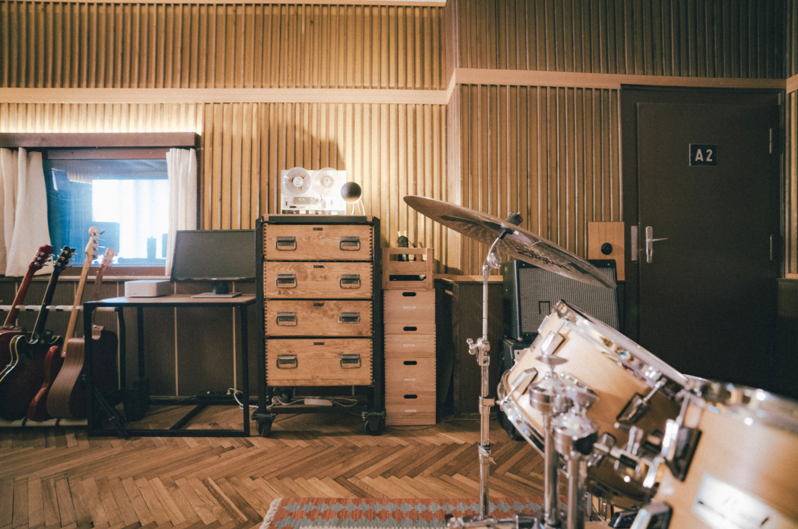 Musik- und Aufnahmeraum mit Gitarren, Schlagzeug, Mischpult, Lautsprecher, Kabel, Holzpaneelen und Fenster.