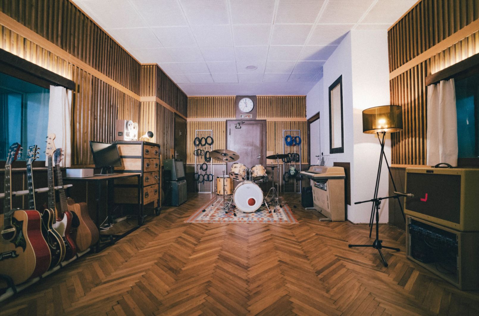 Aufnahme eines Musikproberaums mit verschiedenen Instrumenten, darunter Gitarren auf der linken Seite, ein Schlagzeug in der Mitte, sowie Verstärker und ein Klavier. Der Raum ist mit Holzwänden und einem Teppich im Zentrum ausgestattet.