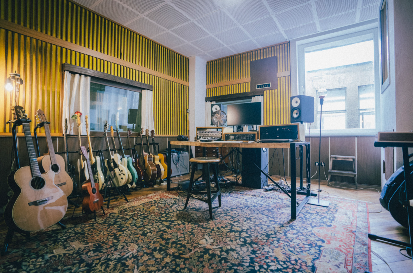 Aufnahme eines Musikstudios mit mehreren Gitarren, einem Computer, Lautsprechern und Musiktechnik auf einem Holztisch, im Raum befinden sich große Fenster, eine Wand mit Holzverkleidung und eine bunte Teppichboden.