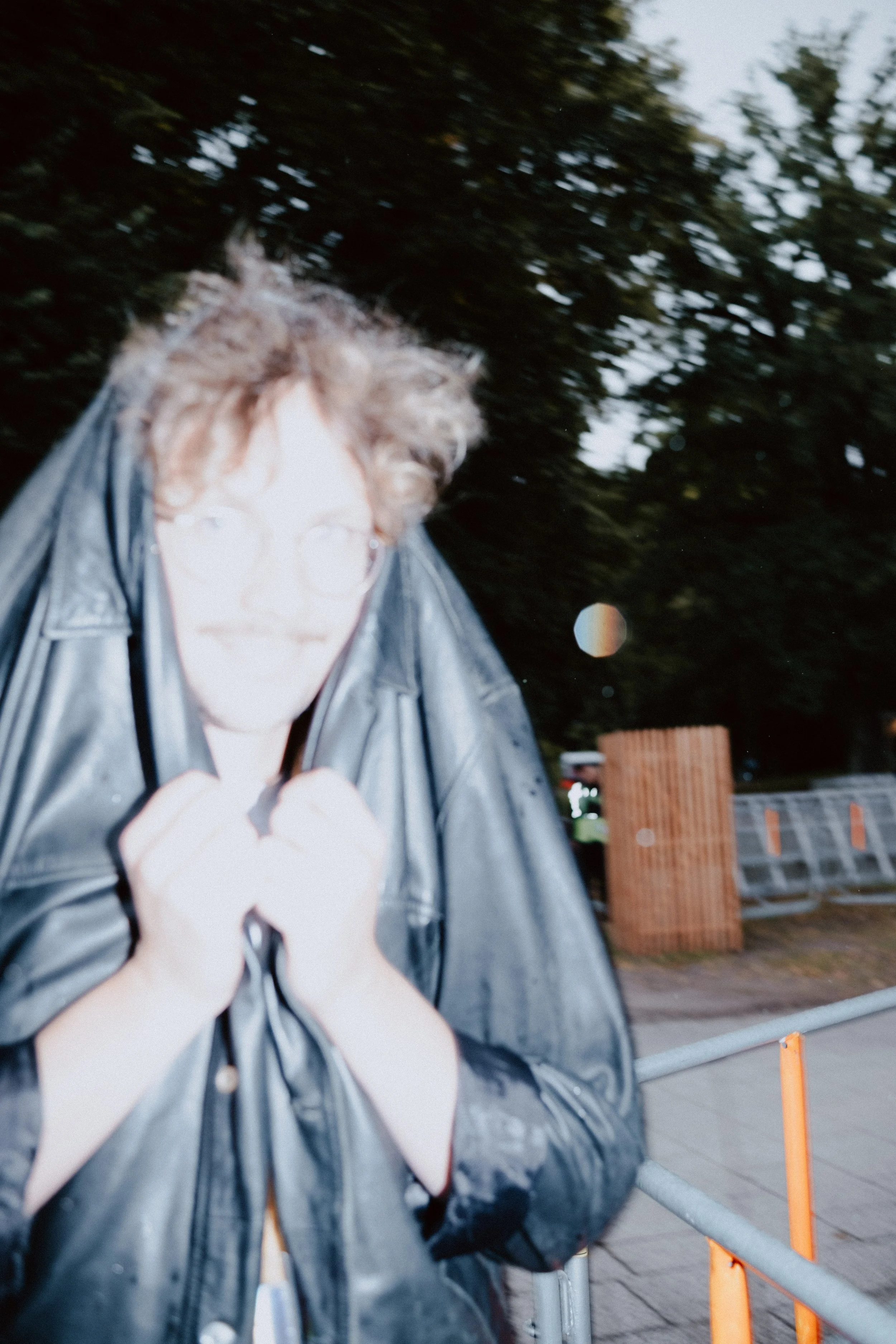 Eine Person mit lockigem Haar und Brille, die eine schwarze Ledermantel trägt, steht draußen vor einer Barriere bei Dämmerung.