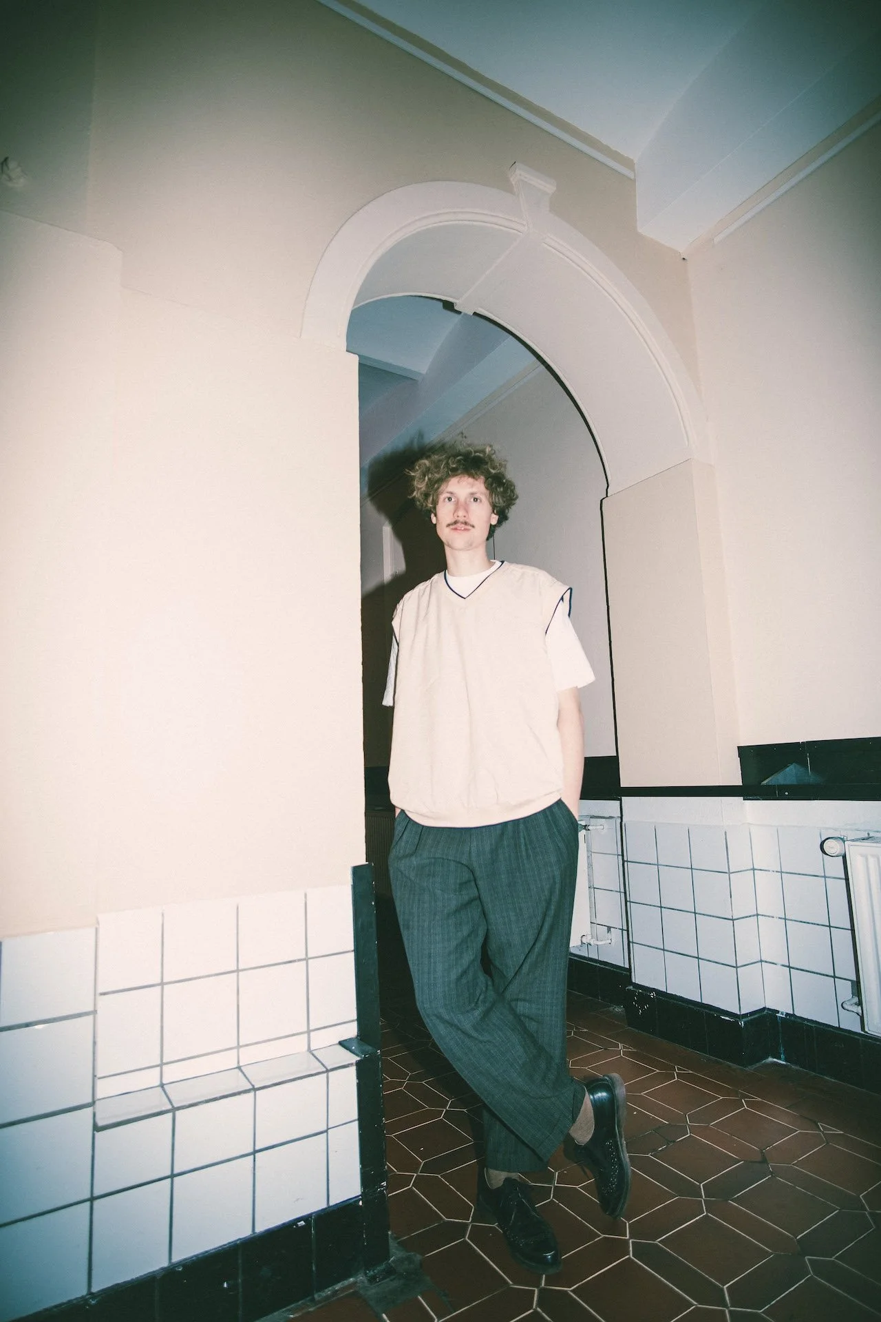 Ein junger Mann mit lockigem Haar, Schnurrbart und legere Kleidung, lehnt entspannt an einer Wand in einem Raum mit weißen Fliesen und einer hellen Wand. Er trägt schwarze Schuhe, eine weiße Weste oder ein Hemd, das aus Hosen herausragt, und schaut direkt in die Kamera.