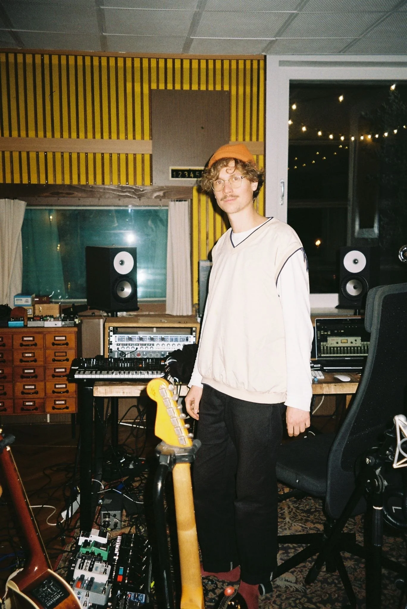 Ein junger Mann in einem Tonstudio mit verschiedenen Musikanlagen, einem Keyboard und einem E-Gitarre. Er trägt eine weiße Weste, eine orange Mütze und hat lockige Haare.