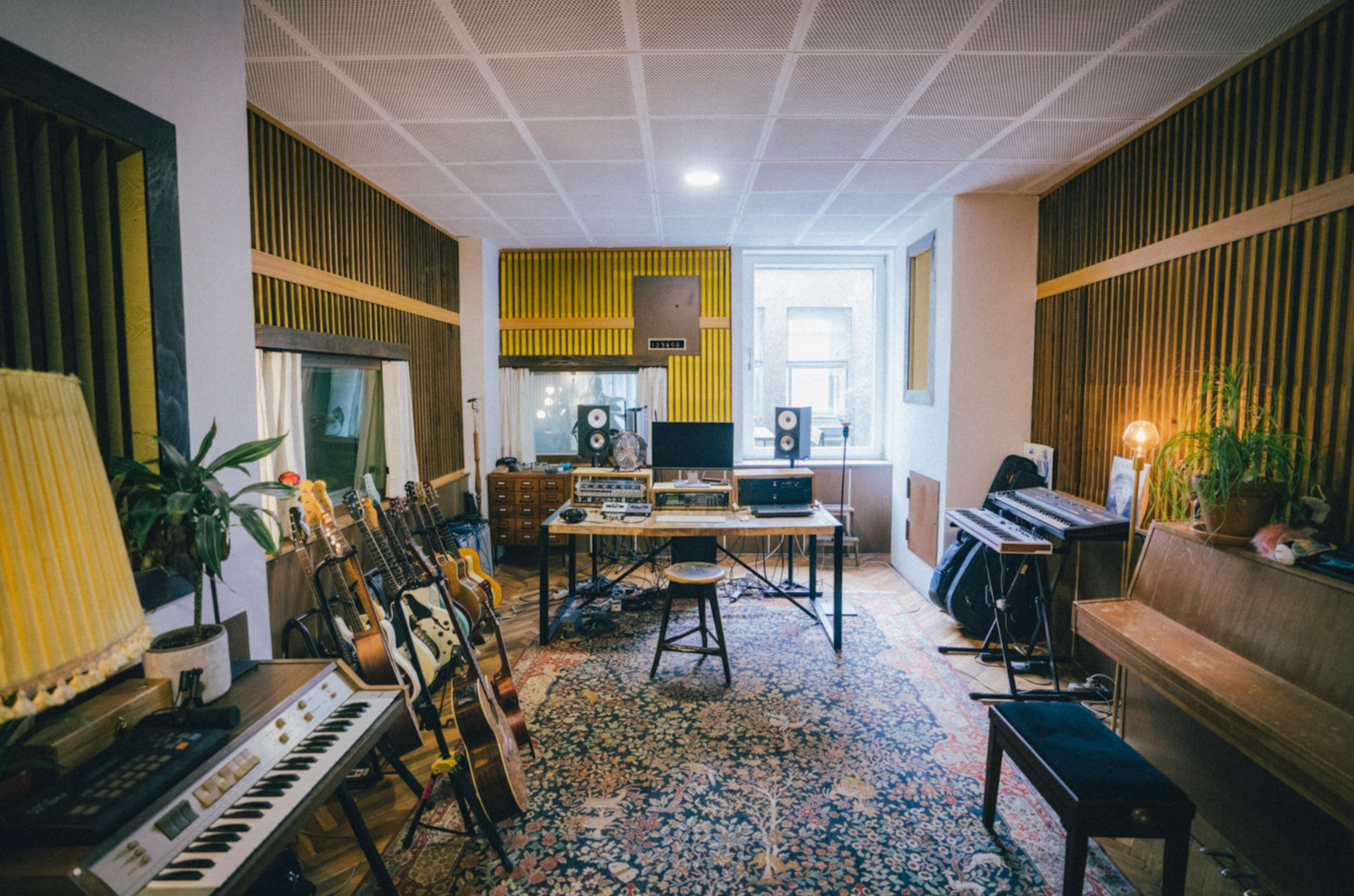 Musikstudio mit Gitarren, elektronischen Instrumenten, einem Keyboard, Lautsprechern, einem Computer, und Recording-Ausrüstung, in einem Raum mit akustischer Wandgestaltung und Teppich.