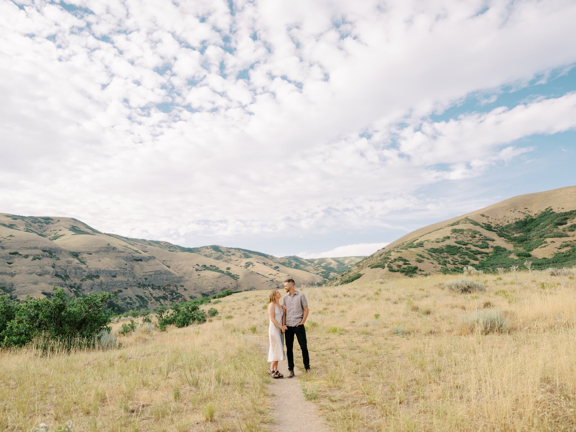 Couple standing on a trail beneath mountains and a cloud-filled summer sky