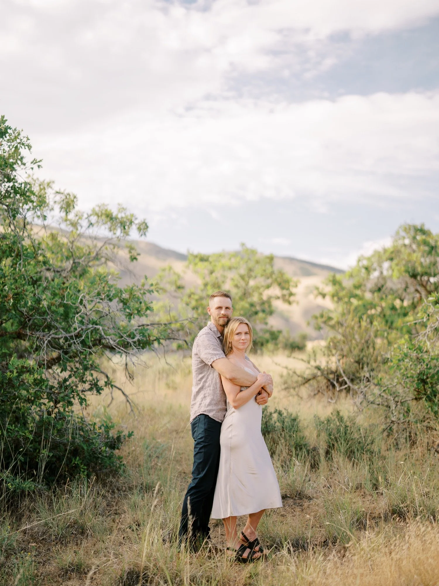 Couple hugging along a grassy trail with trees in the background