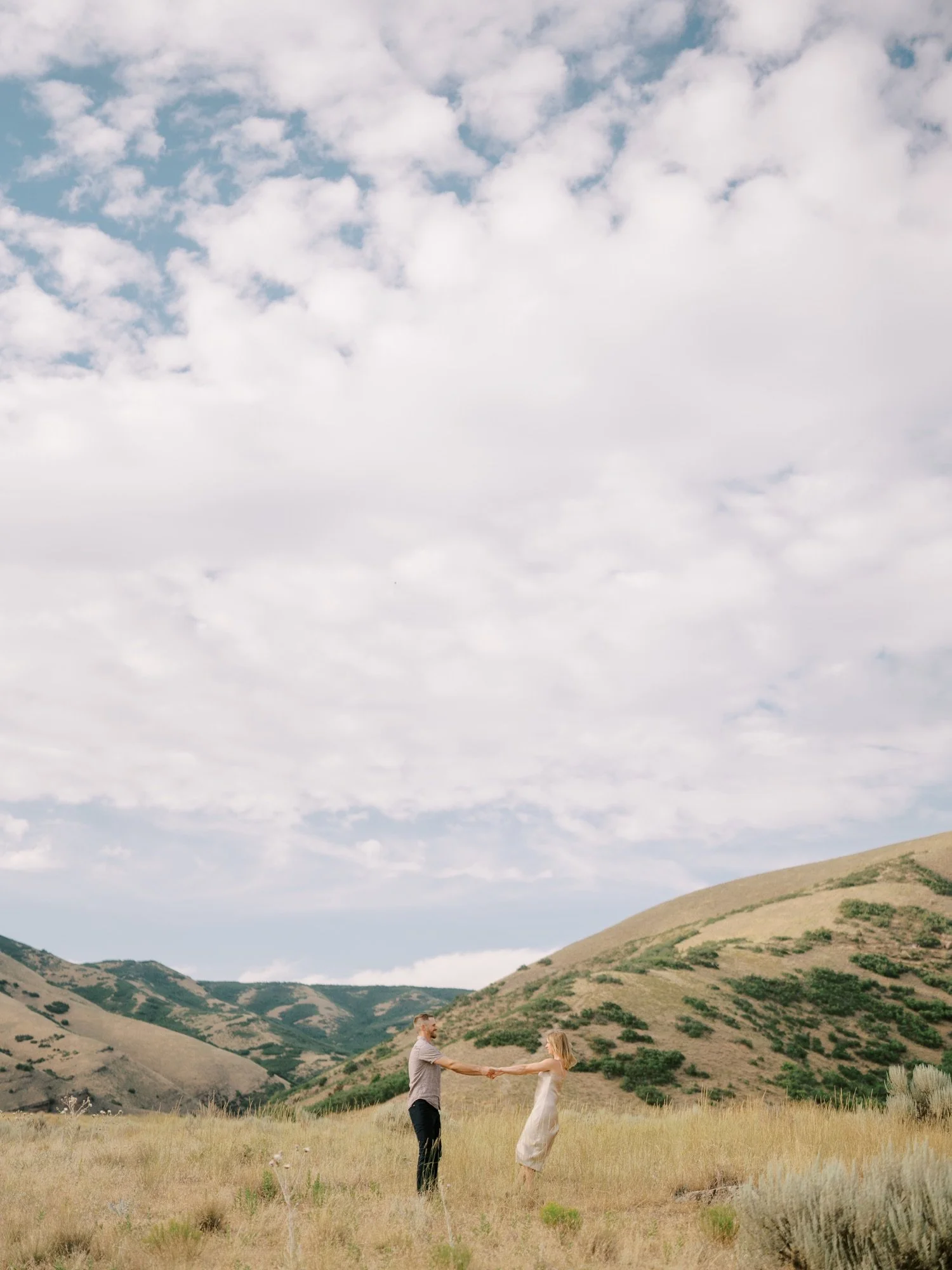 Couple standing in a meadow beneath textured clouds and rolling hills