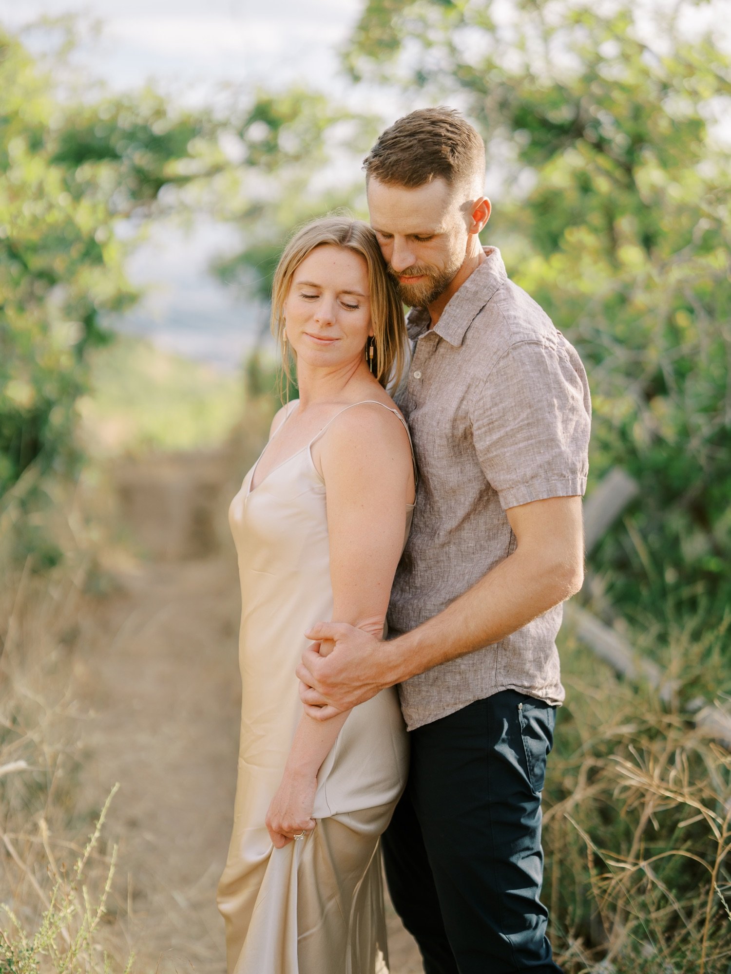 Man kissing his fiancée on the head beside green trees