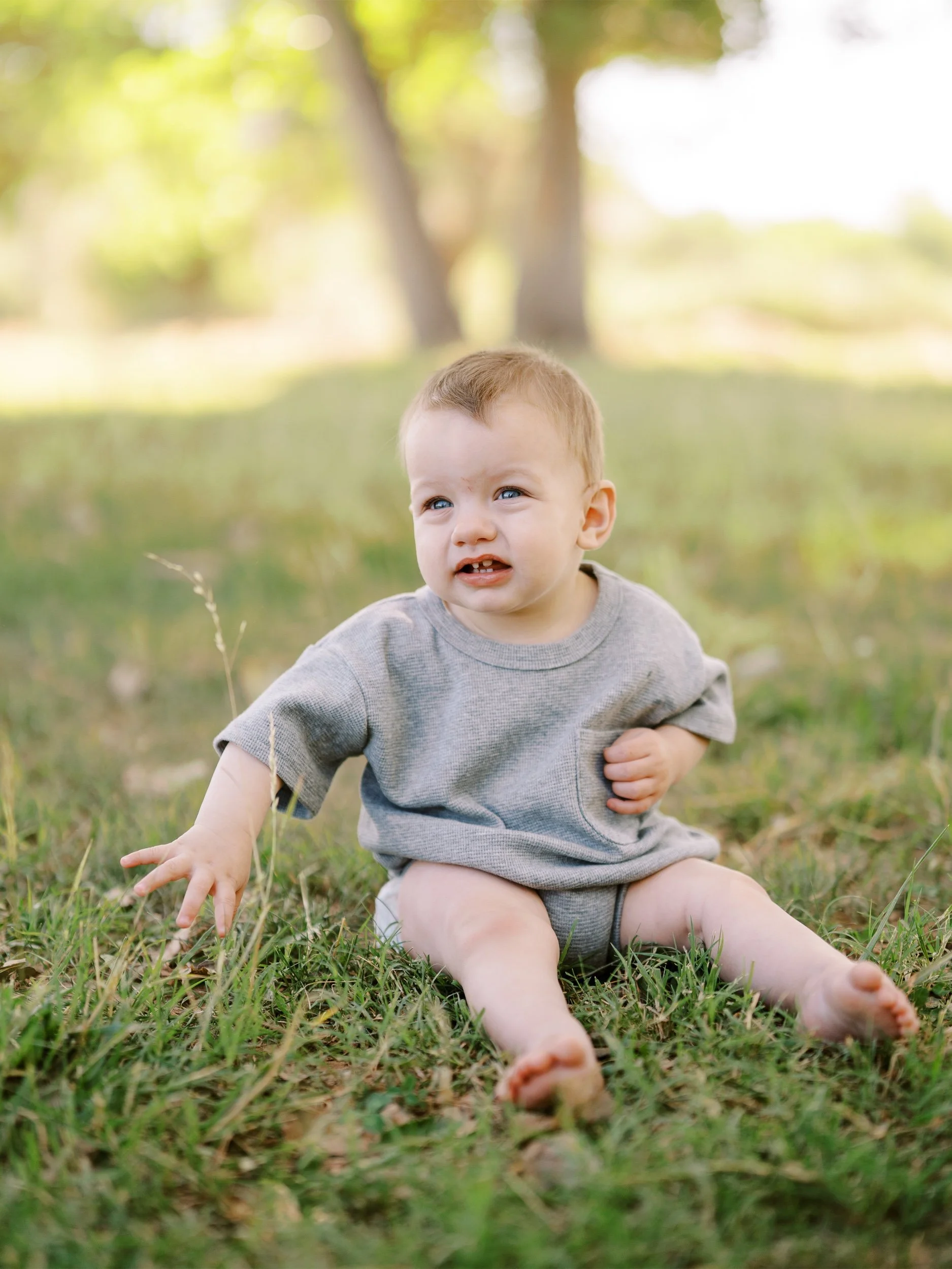 A baby in a gray outfit sitting barefoot on the grass.