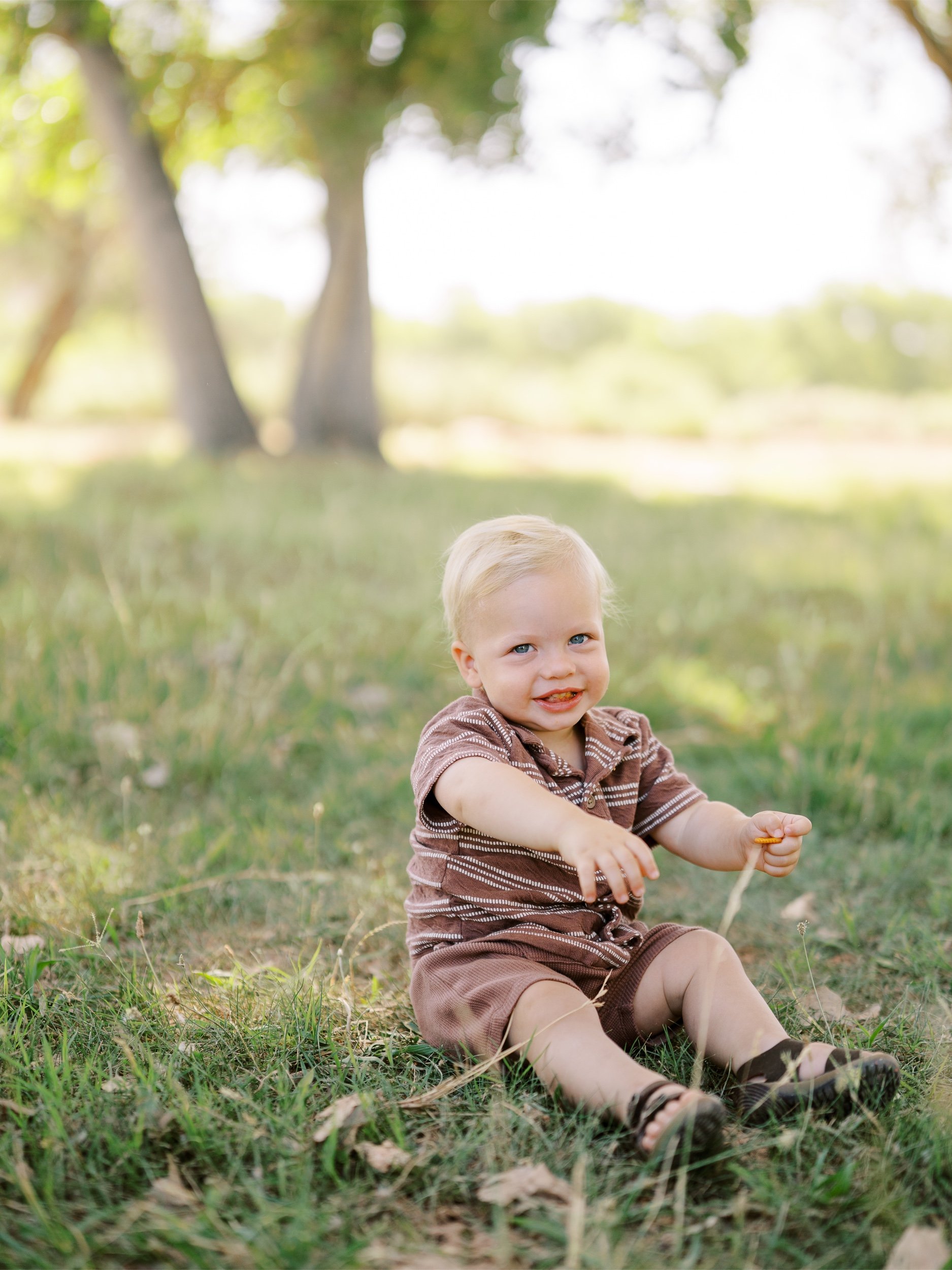 A smiling baby in a brown outfit sitting on the grass.