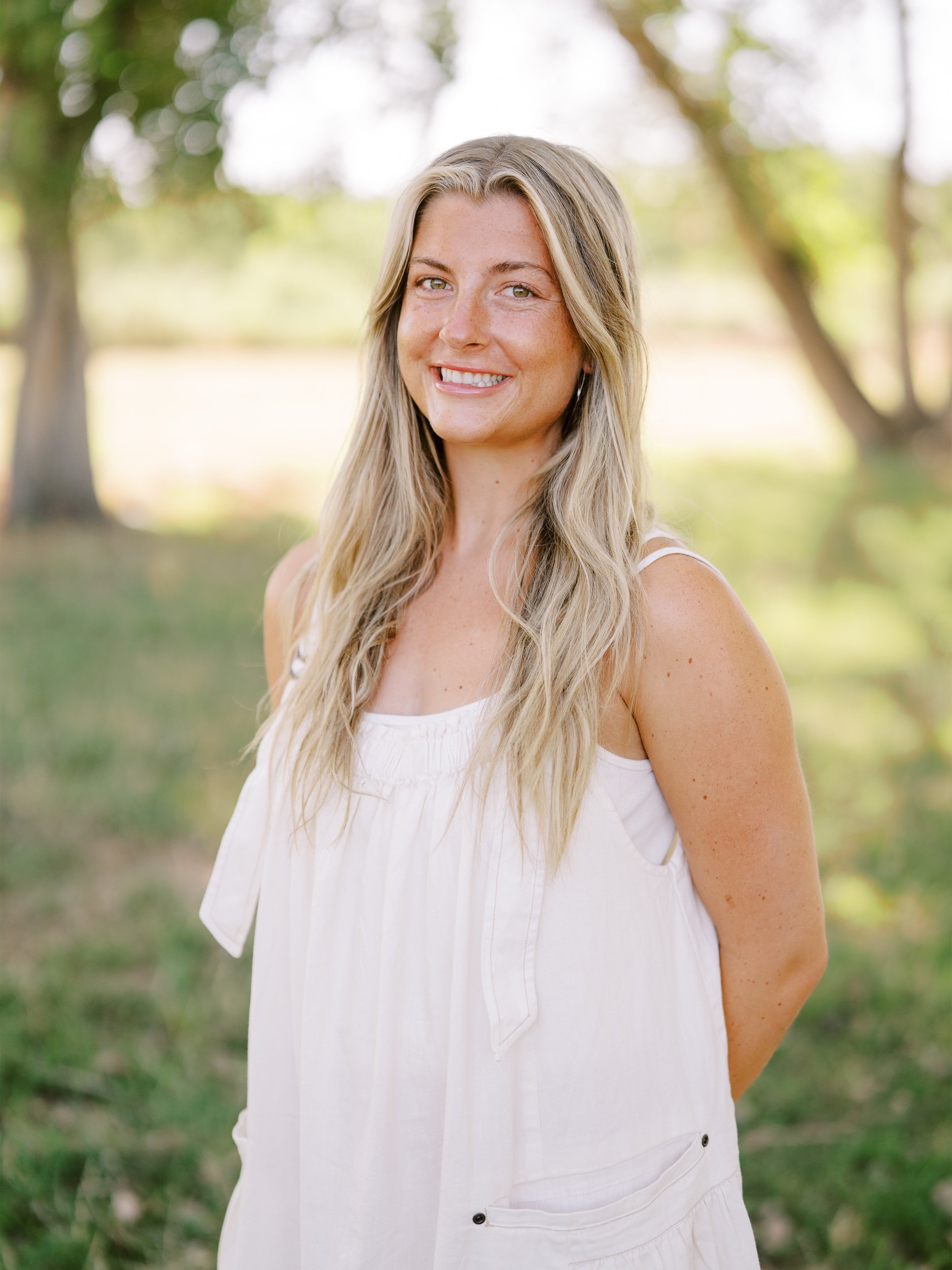 A woman in a cream sleeveless top smiling beneath the trees.