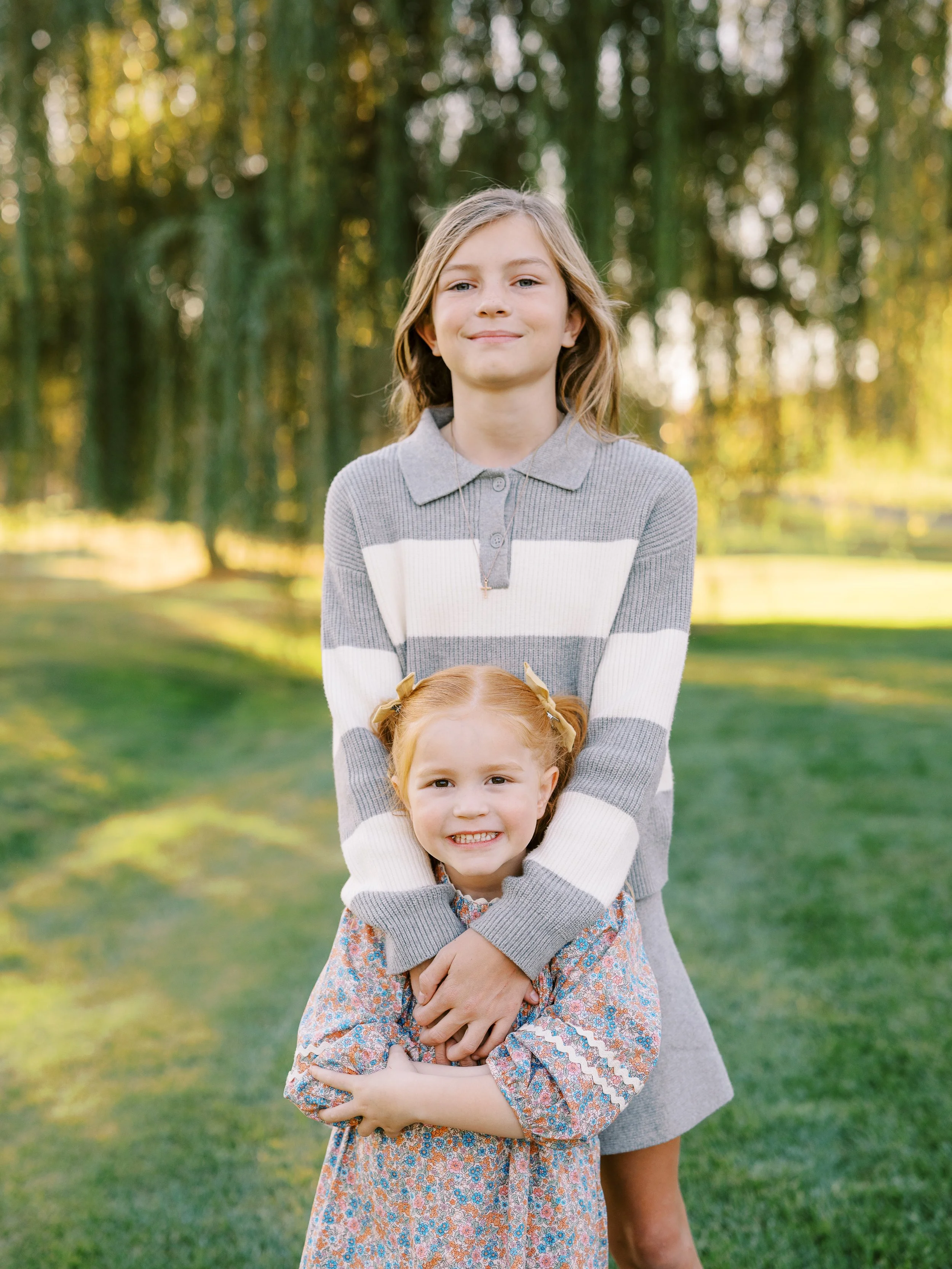 Older sister hugging her younger sister from behind beneath a willow