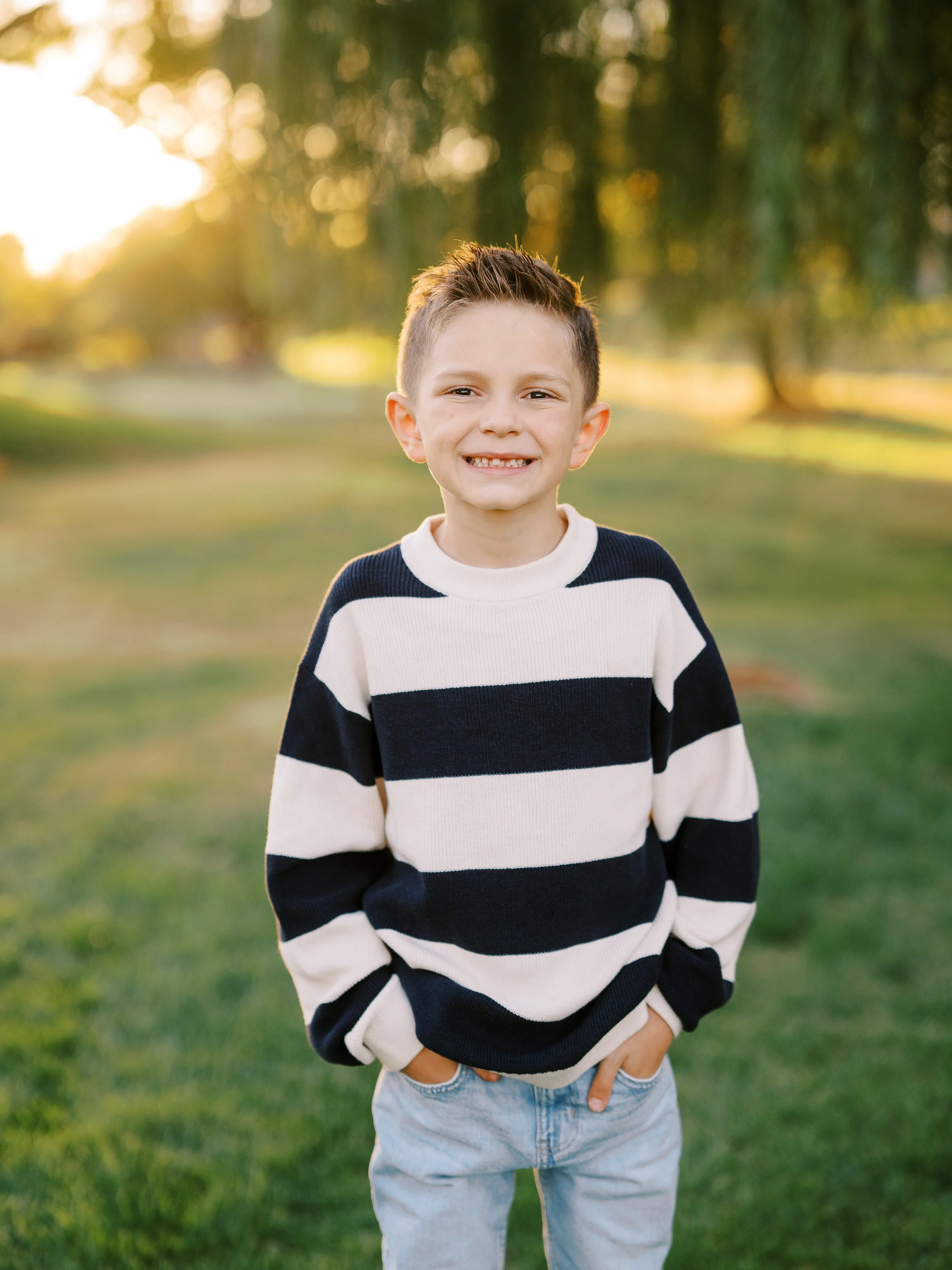 Smiling boy with his hands in his pockets and sunlit trees behind him