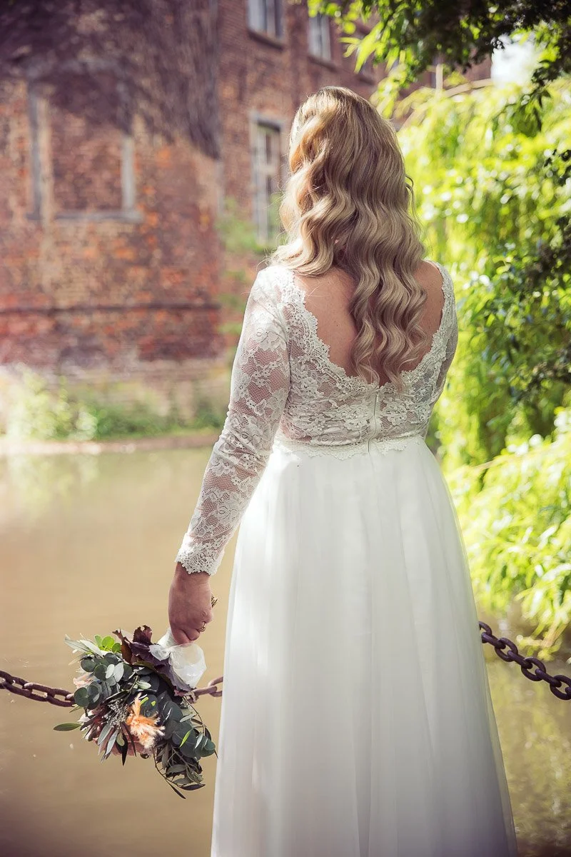 Frau in Hochzeitskleid steht an einem Fluss, umgeben von grünen Bäumen, hält einen Brautstrauß in der Hand und schaut weg, mit Backsteinmauer im Hintergrund.