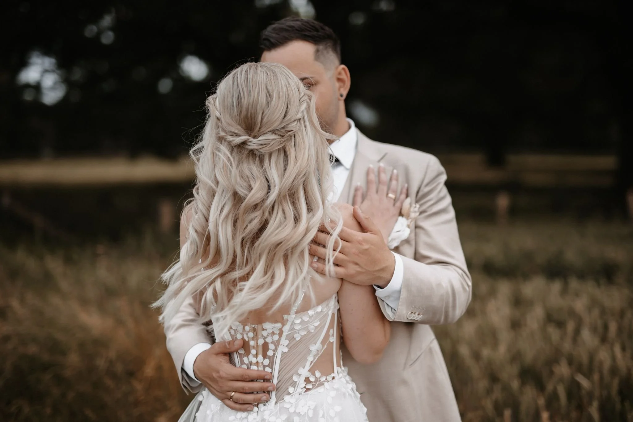 Ein Brautpaar beim Tanzen im Freien, die Frau trägt ein weißes Hochzeitskleid mit Blumenmuster, der Mann in einem hellen Anzug. Sie umarmen sich eng in einer natürlichen Umgebung.