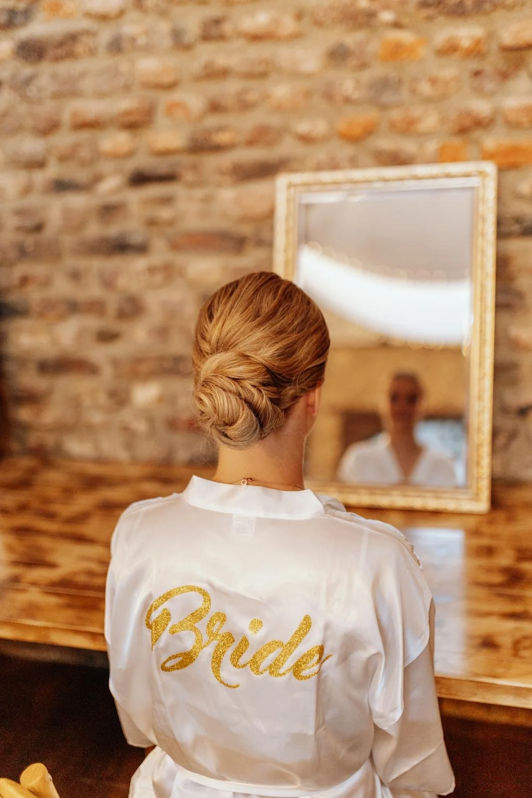 Braut mit aufwendig gestyltem Haar sieht in einem Spiegel, während sie eine weiße Robe mit goldenem Aufdruck 'Bride' trägt, in einem Raum mit Ziegelsteinwand.