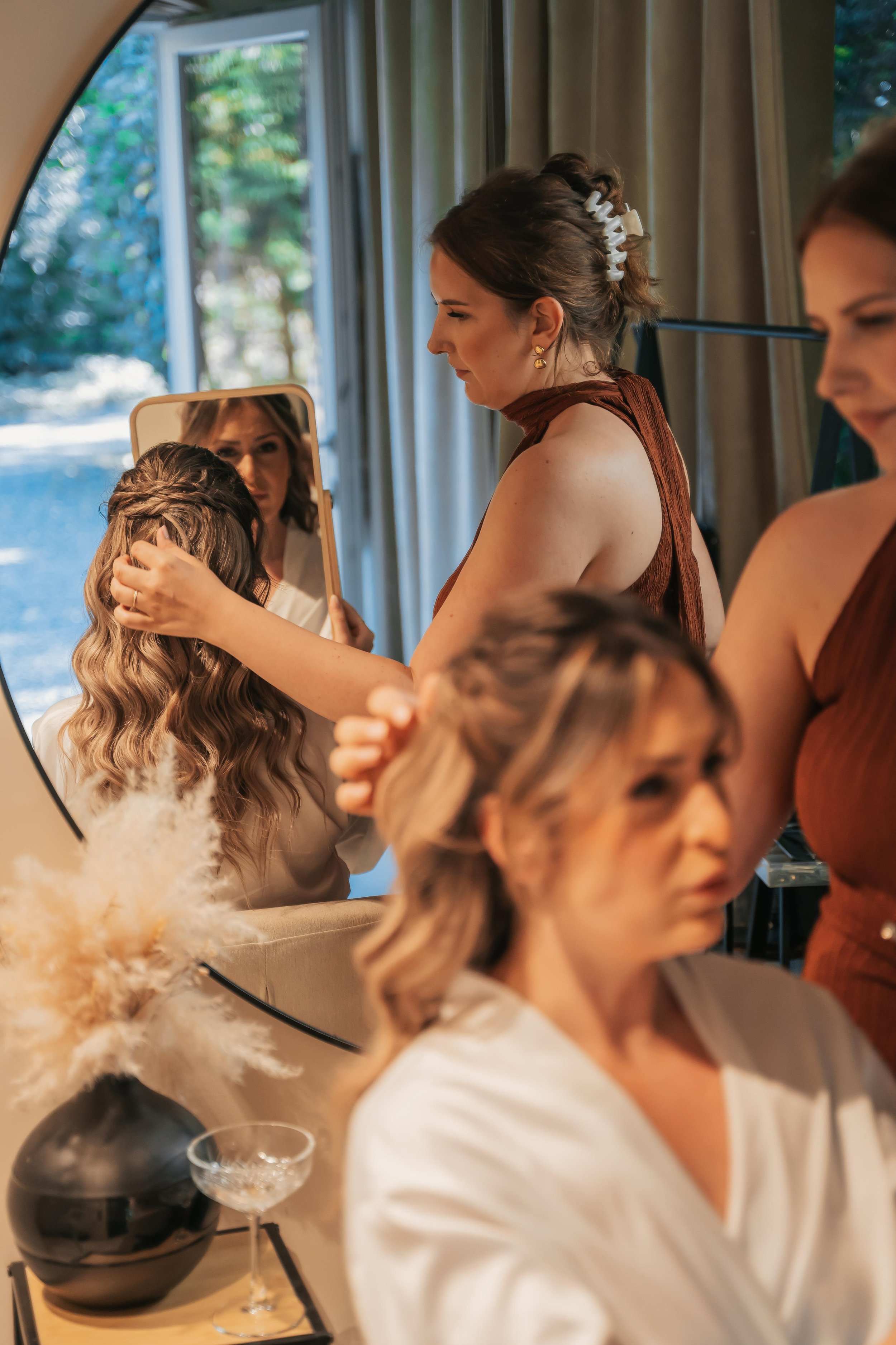Eine Frau beim Frisieren, im Spiegel sichtbar, mit anderen Frauen im Hintergrund bei einer Hochzeit oder besonderen Gelegenheit, im Raum mit großen Fenstern und Vorhängen.