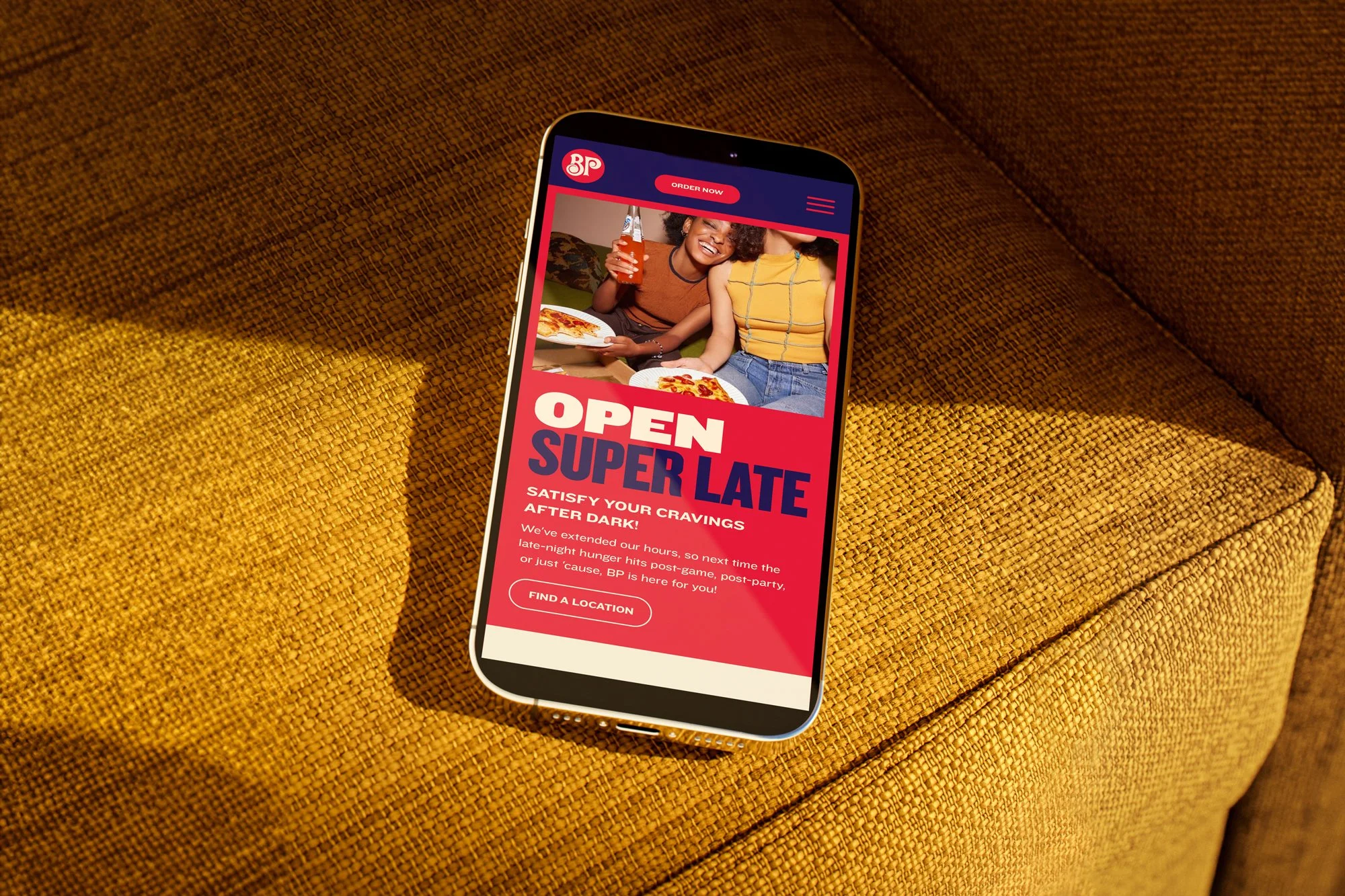 Smartphone on yellow textured couch displaying a Burger King online menu advertising late-night food service with a photo of two women enjoying pizza and drinks.