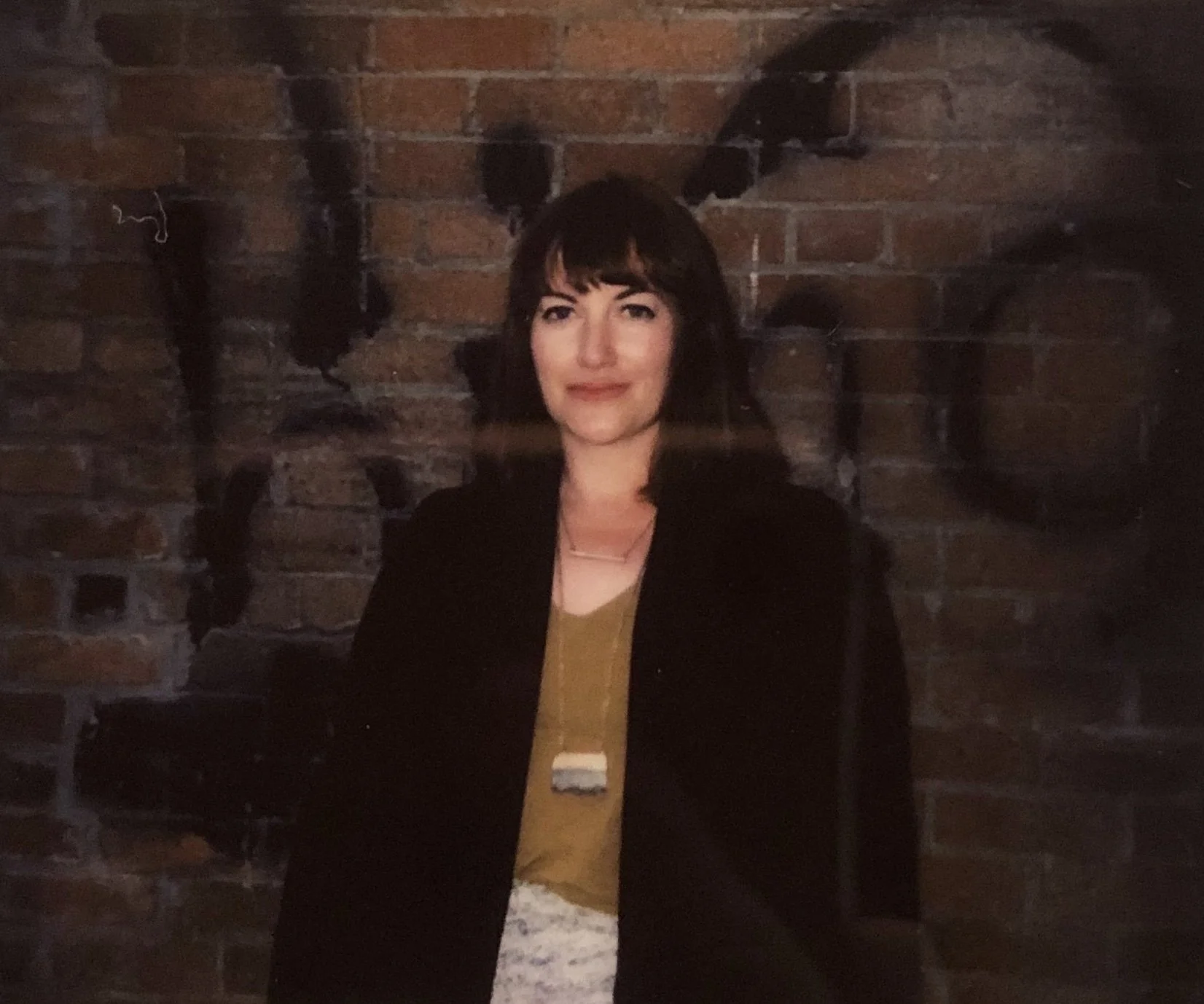 A woman with dark hair and bangs standing in front of a brick wall with black abstract paint designs.