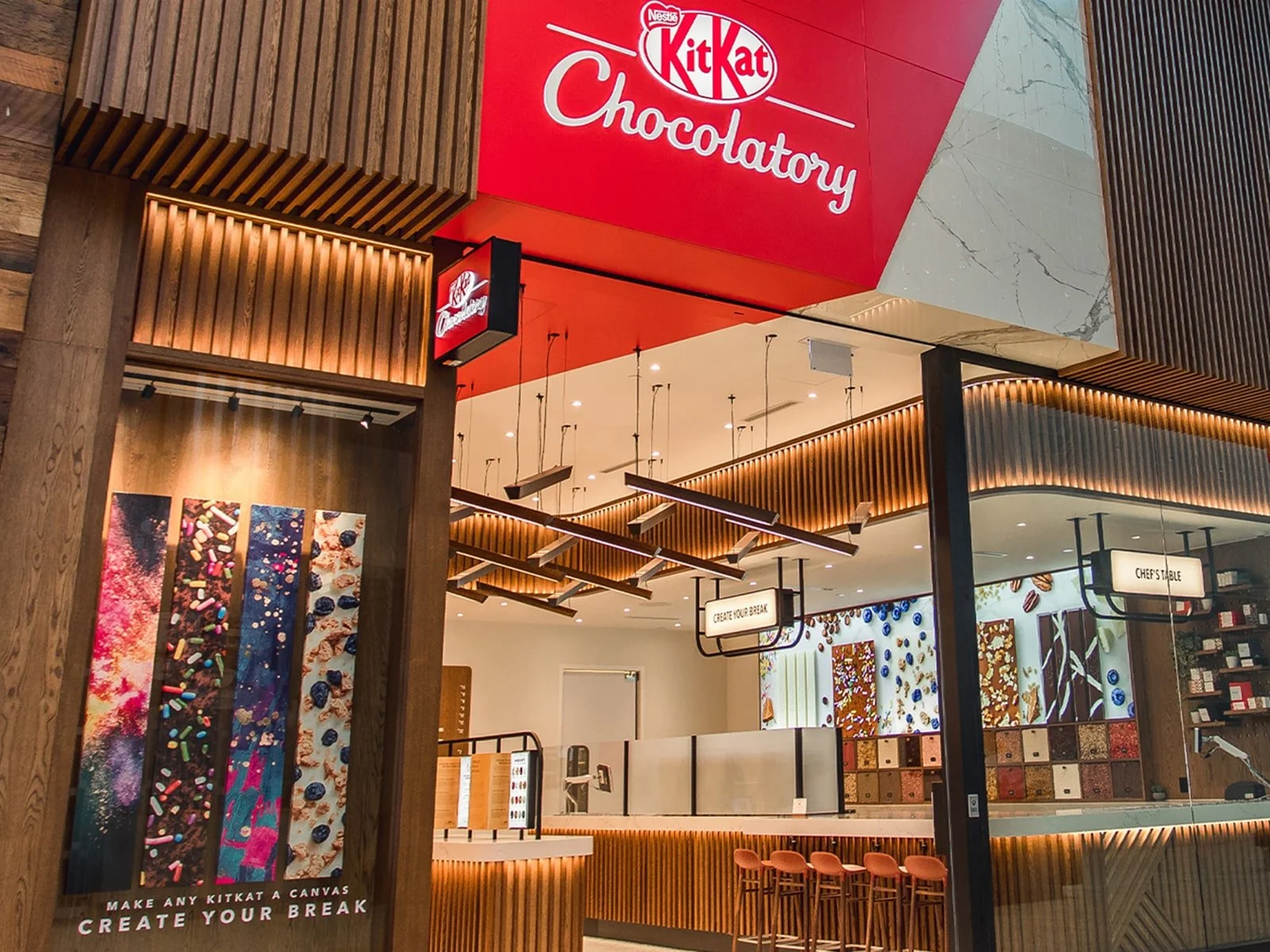 Inside a KitKat Chocolatory store, with a modern design featuring wood accents and colorful artwork on the walls, and a counter with bar stools for creating personalized chocolates.